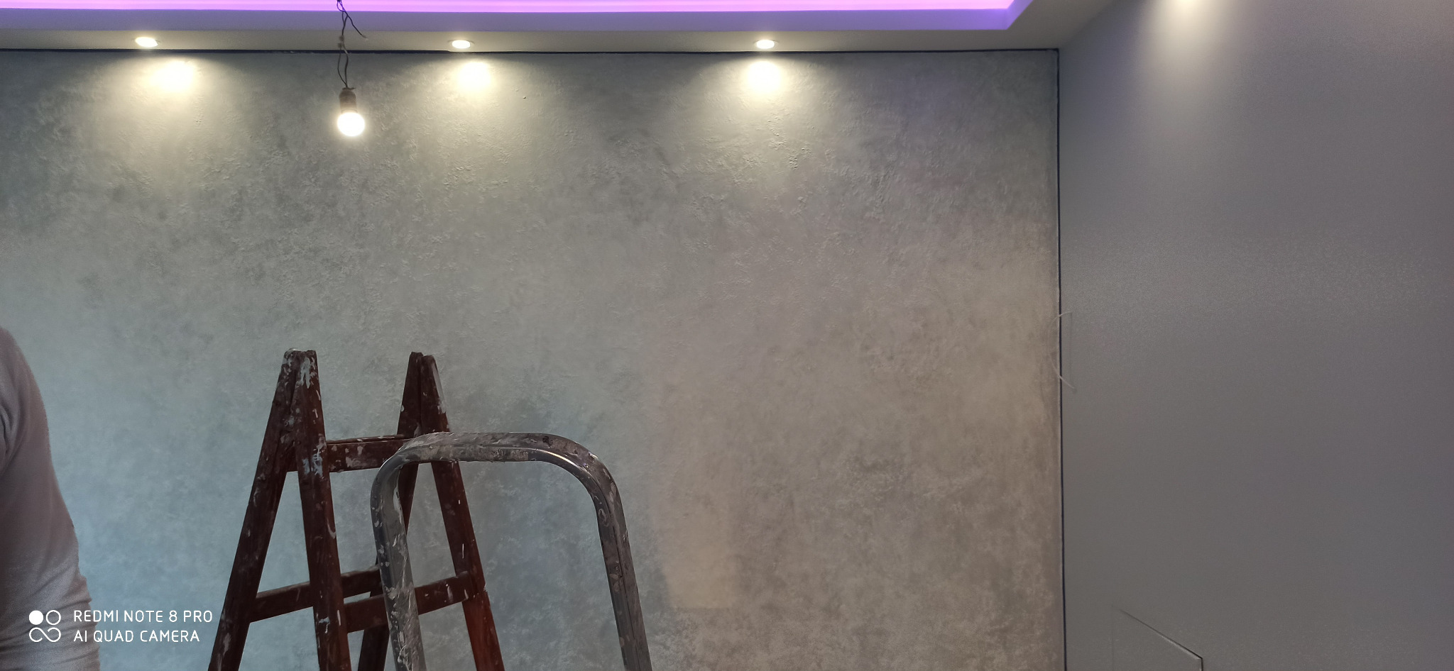 Ściana z efektem dekoracyjnym w odcieniach szarości, oświetlona punktowymi lampami sufitowymi i listwą LED, z widoczną częścią drabiny malarskiej.