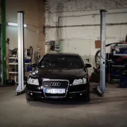 Czarny samochód Audi A6 na podnośniku dwukolumnowym w warsztacie z białymi, ceglanymi ścianami i betonową podłogą.