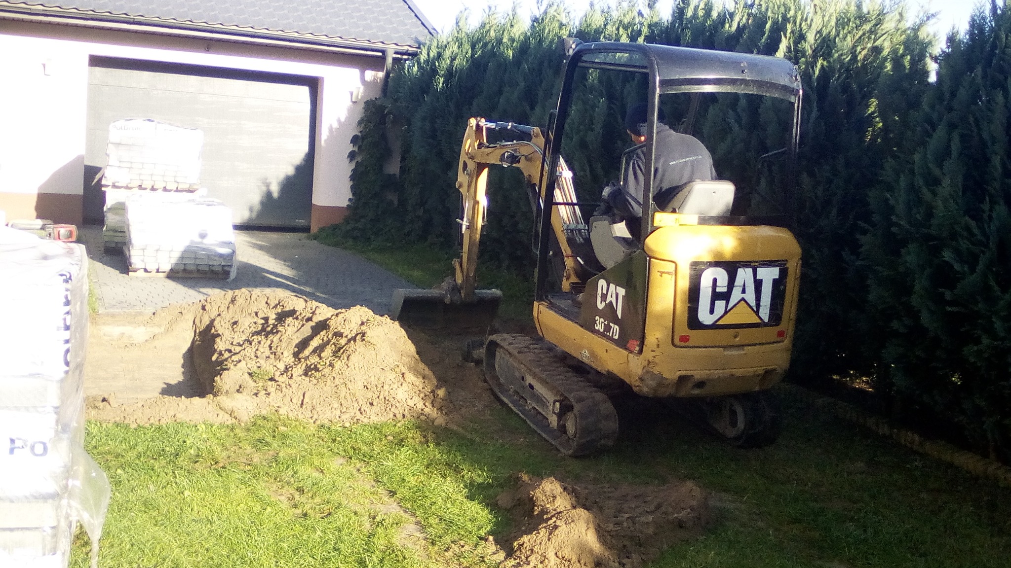 Żółta mini koparka CAT 301.7D wykopuje rów na trawniku, obok zapakowane palety z kostką brukową i garaż.