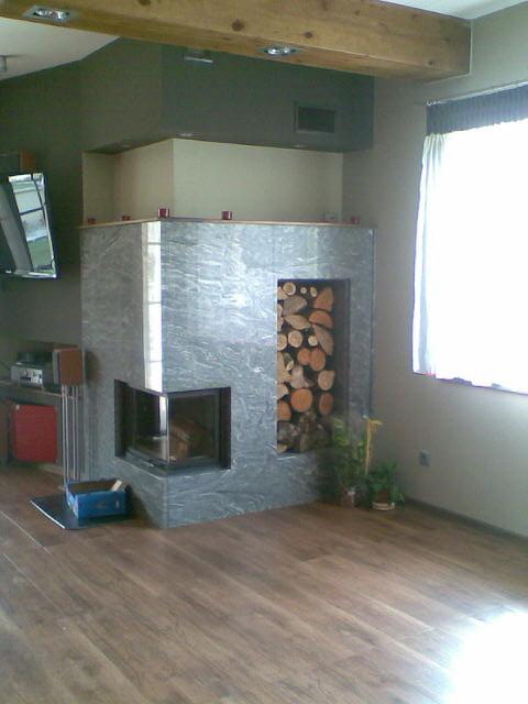 Szary kominek z marmurowym wykończeniem, z wnęką na drewno, na tle drewnianej podłogi i fragmentu salonu z telewizorem i belką stropową.