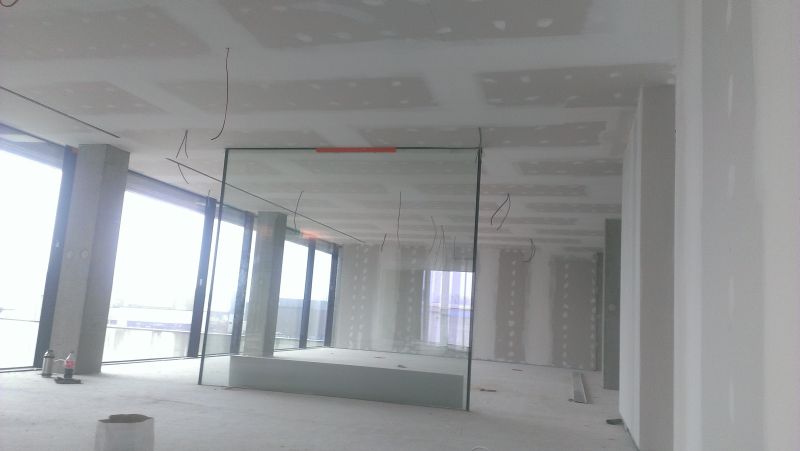 Wnętrze w trakcie remontu: ściany pokryte gładzią gipsową, widoczne kable zwisające z sufitu, szklana ścianka działowa, duże okna z betonowymi filarami.