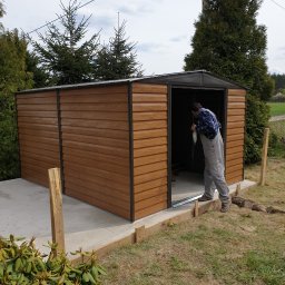 EDPOL USŁUGI REMONTOWE EDWARD SZUBA - Montaż brązowego, metalowego domku narzędziowego na betonowej wylewce. Mężczyzna w szarym kombinezonie wchodzi do środka, trzymając w ręku maseczkę. Widoczne drewniane elementy szalunku. W tle drzewa.