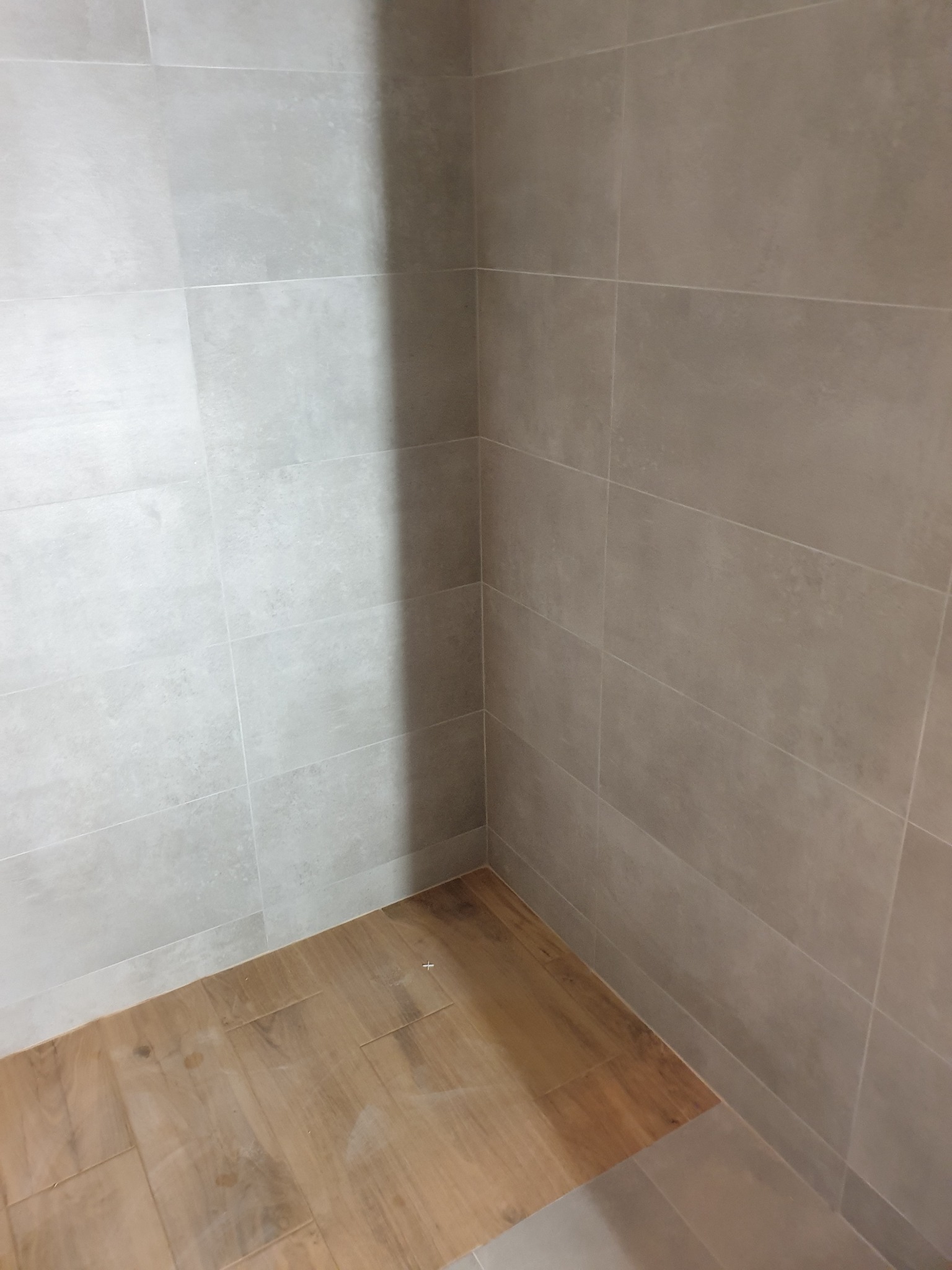 Narożnik łazienki wyłożony szarymi, prostokątnymi płytkami ściennymi i podłogą z płytek imitujących drewno, widoczny cień na ścianie.