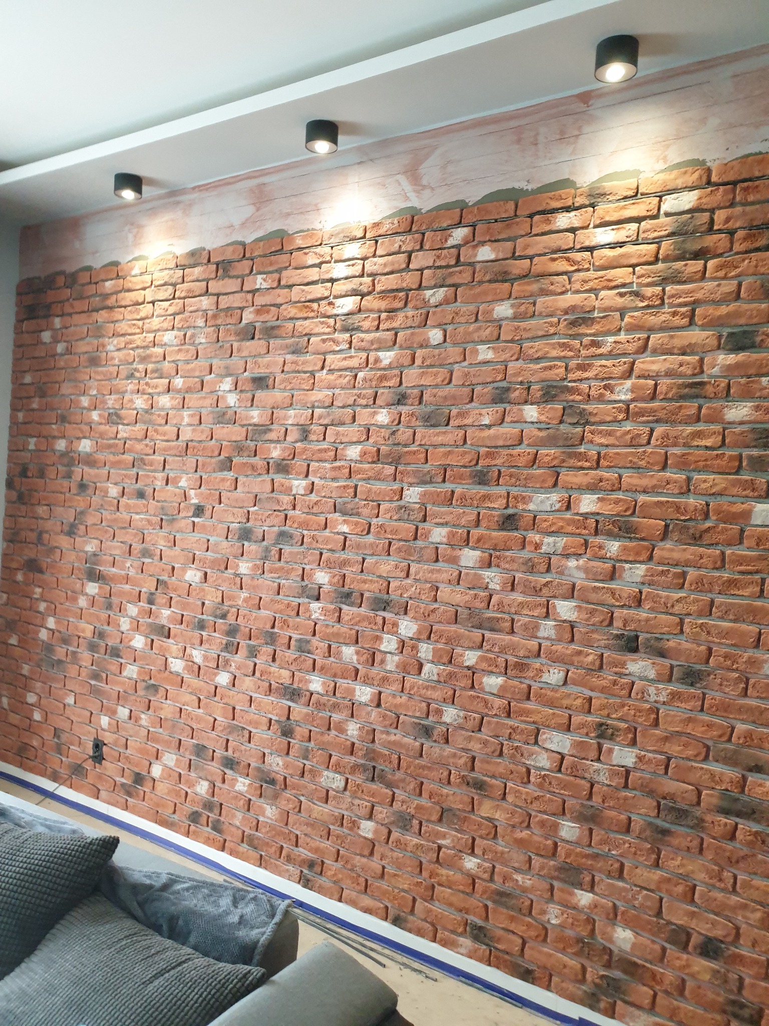 Dekoracyjna ściana z cegły w stylu loft w Łodzi, oświetlona trzema punktowymi lampami sufitowymi, widoczny fragment sofy z poduszkami.