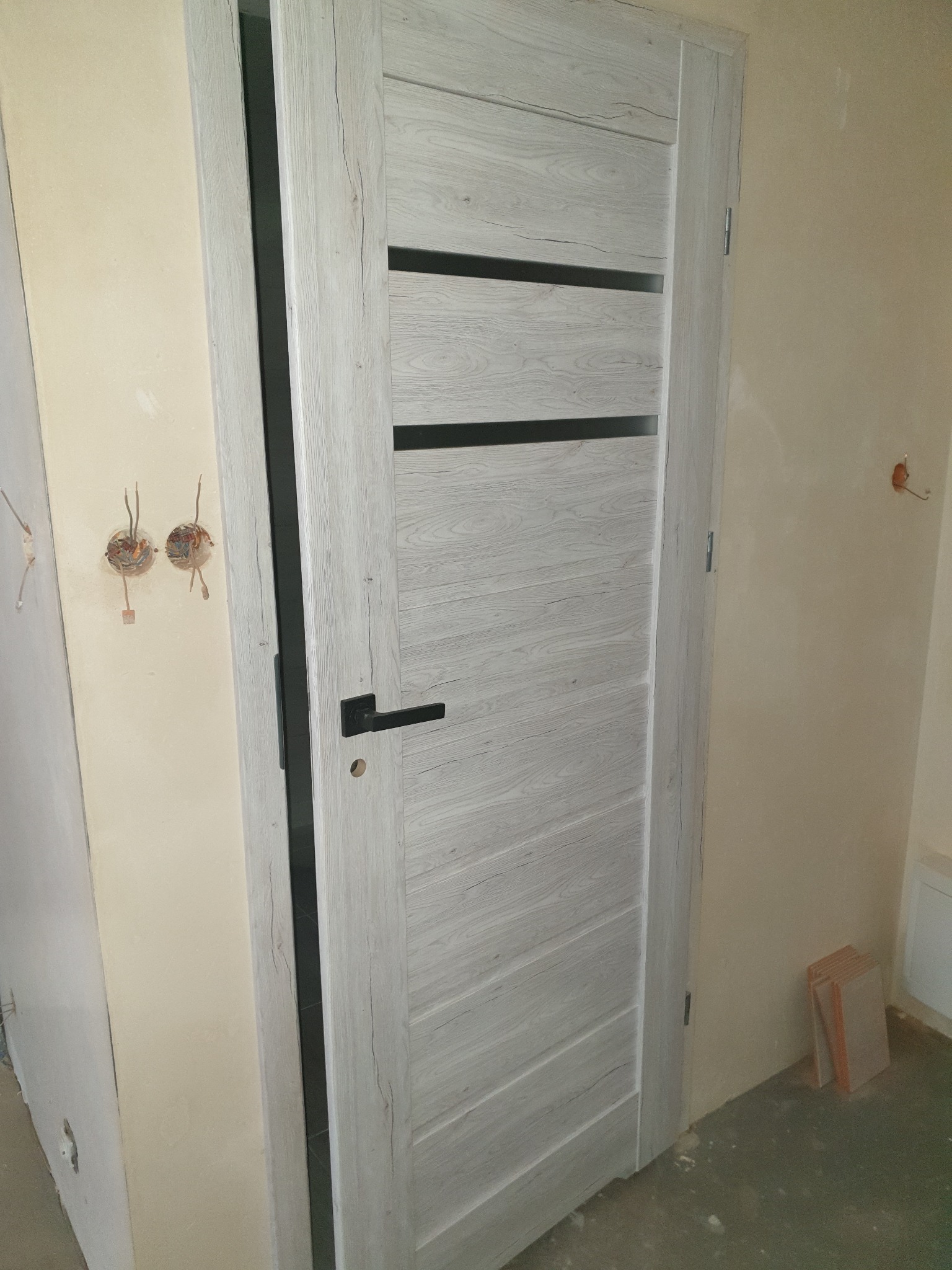 Nowe, białe drzwi wewnętrzne z poziomymi, czarnymi wstawkami, zamontowane w surowym wnętrzu z widocznymi śladami prac wykończeniowych na ścianach i wystającymi kablami elektrycznymi.