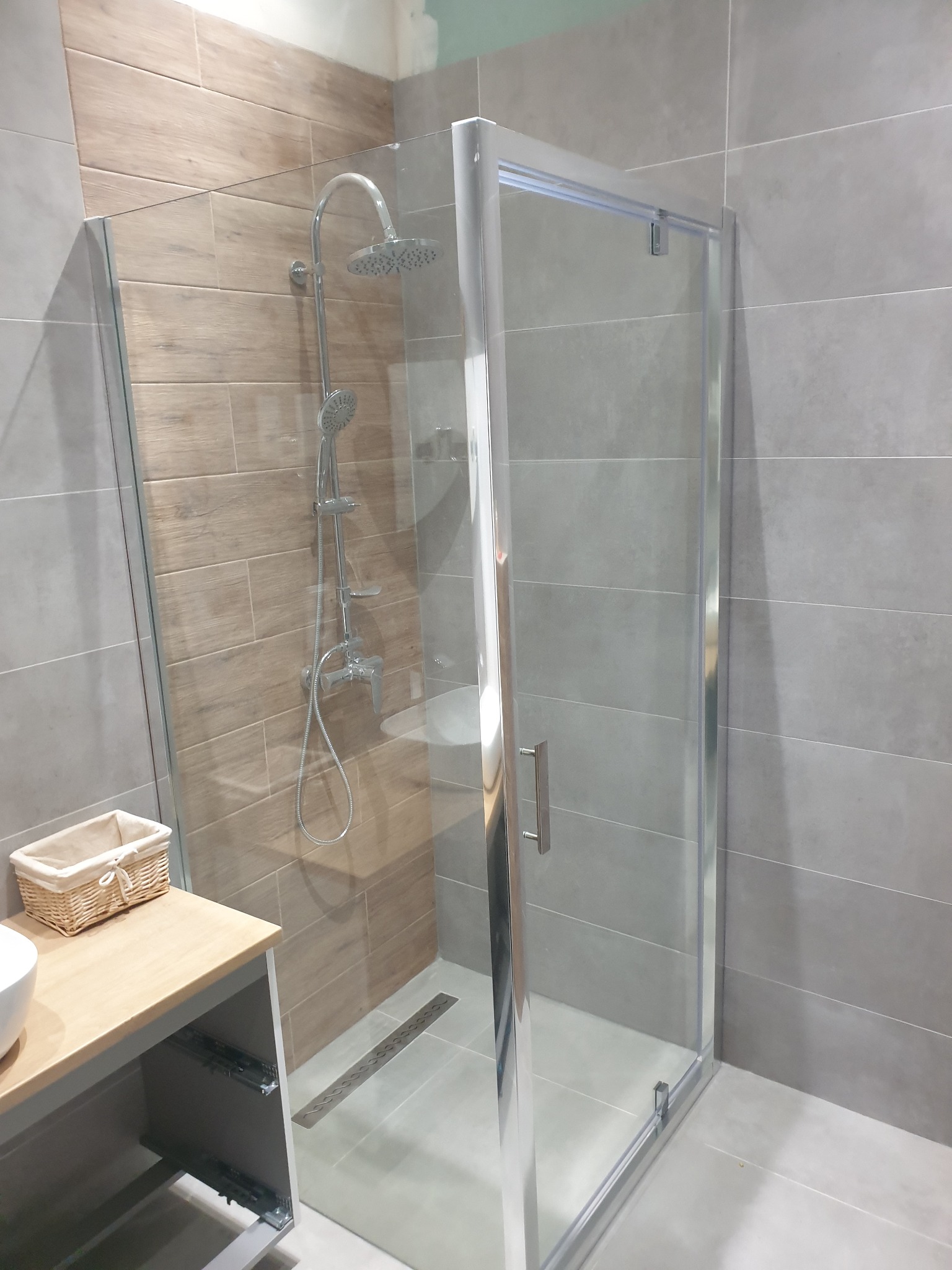 Nowoczesna kabina prysznicowa z przesuwnymi drzwiami, wyłożona płytkami imitującymi drewno na jednej ścianie i szarymi płytkami na pozostałych, z widocznym odpływem liniowym i szafką pod umywalkę...