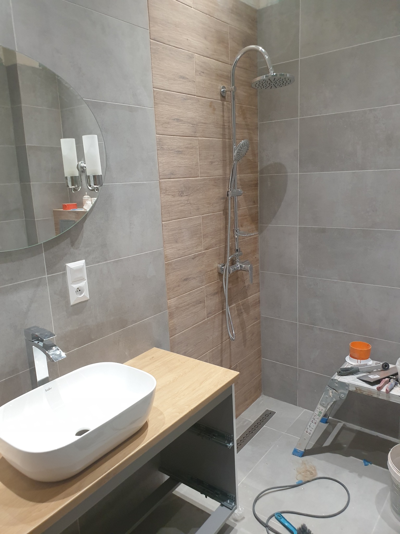 Nowoczesna łazienka w szaro-beżowej kolorystyce z owalną umywalką nablatową, chromowaną armaturą i deszczownicą, częściowo wykończona, widoczny drabinkowy podest z narzędziami i akcesoriami...