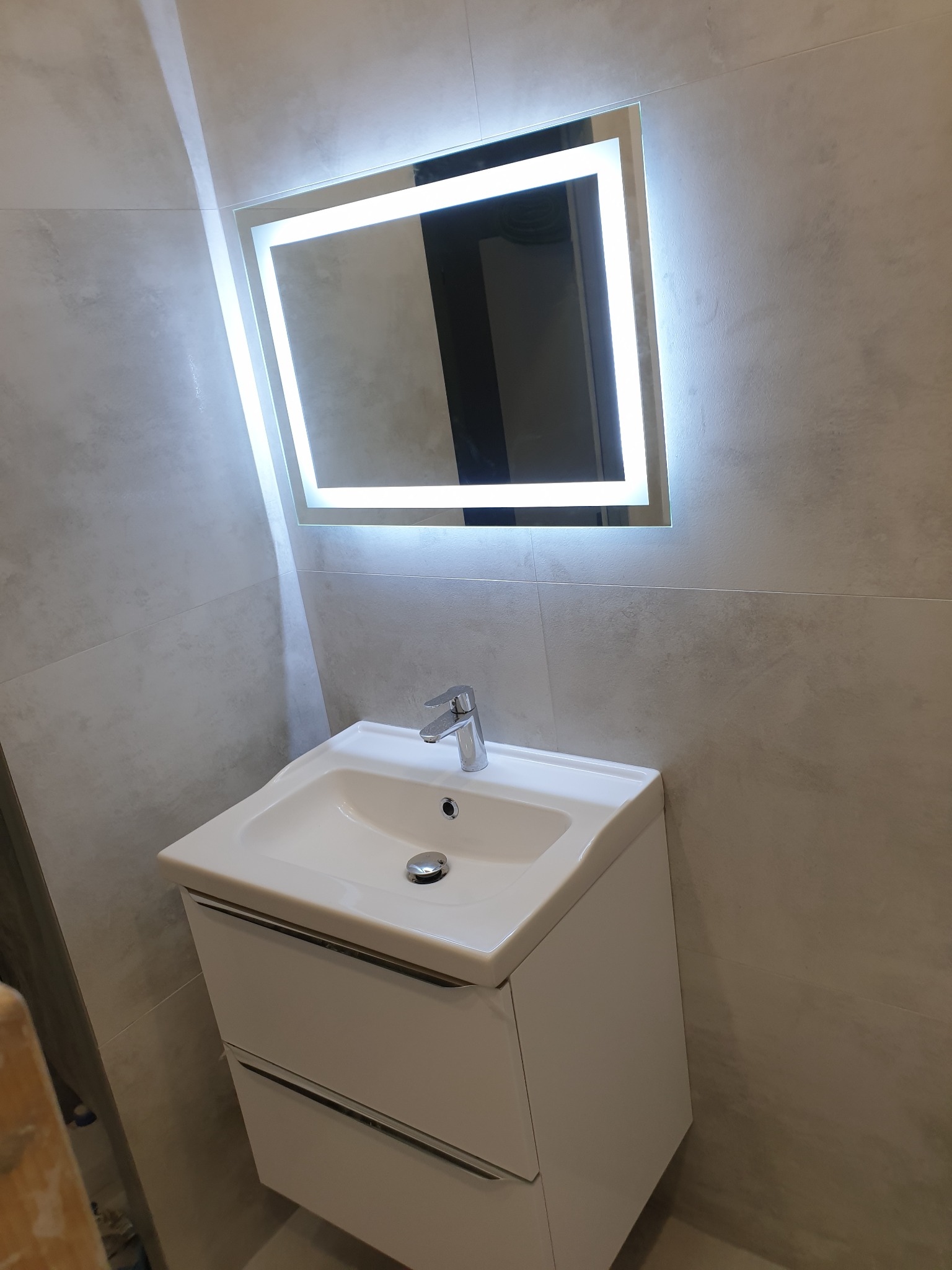 Umywalka z szafką w nowoczesnej łazience, podświetlane lustro LED na szarej płytce, widoczny fragment drabiny malarskiej w lewym dolnym rogu.