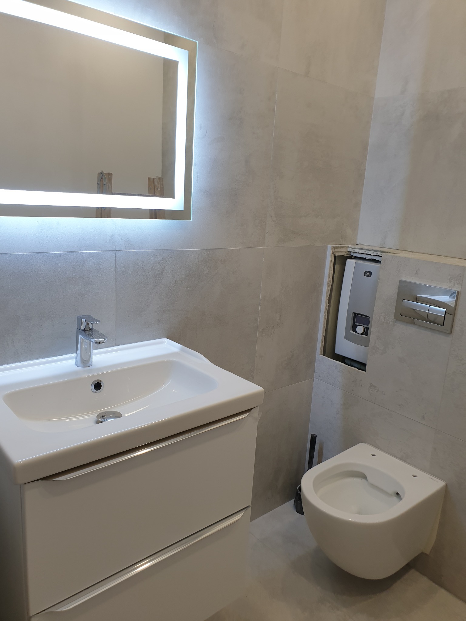 Nowoczesna łazienka w szarościach z podwieszaną toaletą, umywalką z szafką i lustrem z podświetleniem LED, widoczny fragment instalacji w ścianie