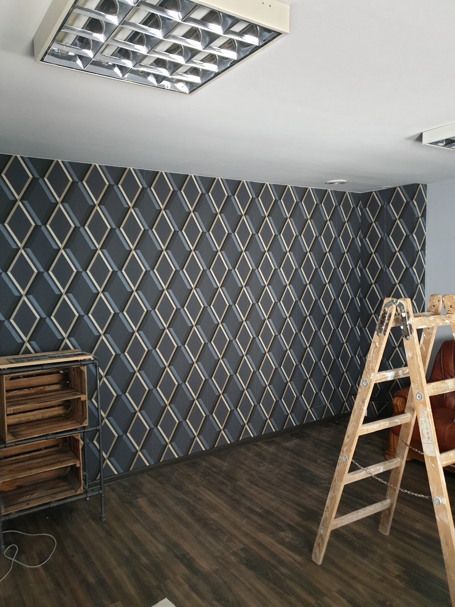 Pokój z nową, geometryczną tapetą w odcieniach szarości i złota, drewniana drabina oparta o ścianę, podłoga z paneli imitujących drewno, metalowy regał z drewnianymi skrzynkami.