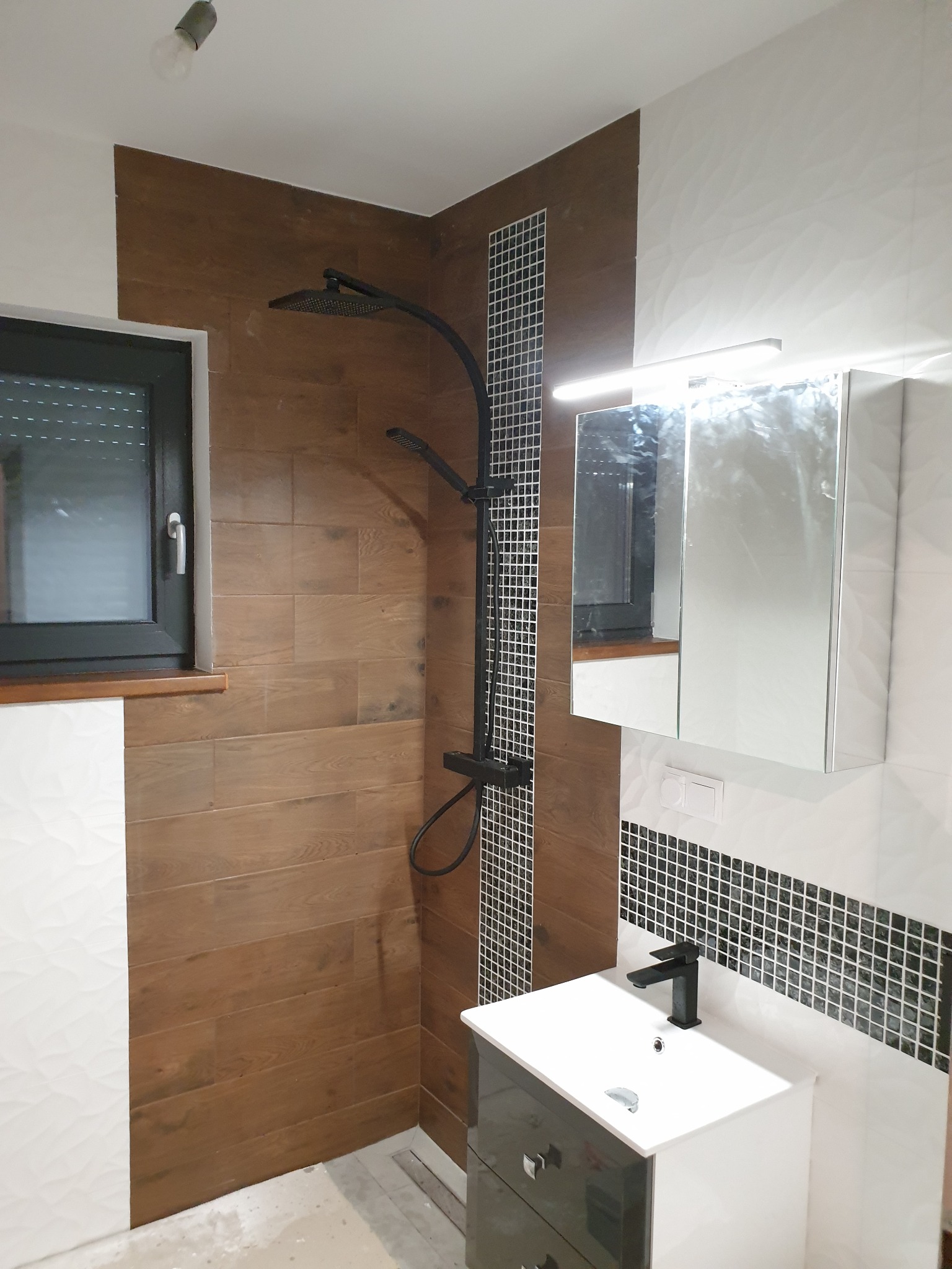 Nowoczesna łazienka z brązowymi płytkami imitującymi drewno, czarną armaturą, szafką z lustrem i dekoracyjnym pasem mozaiki.