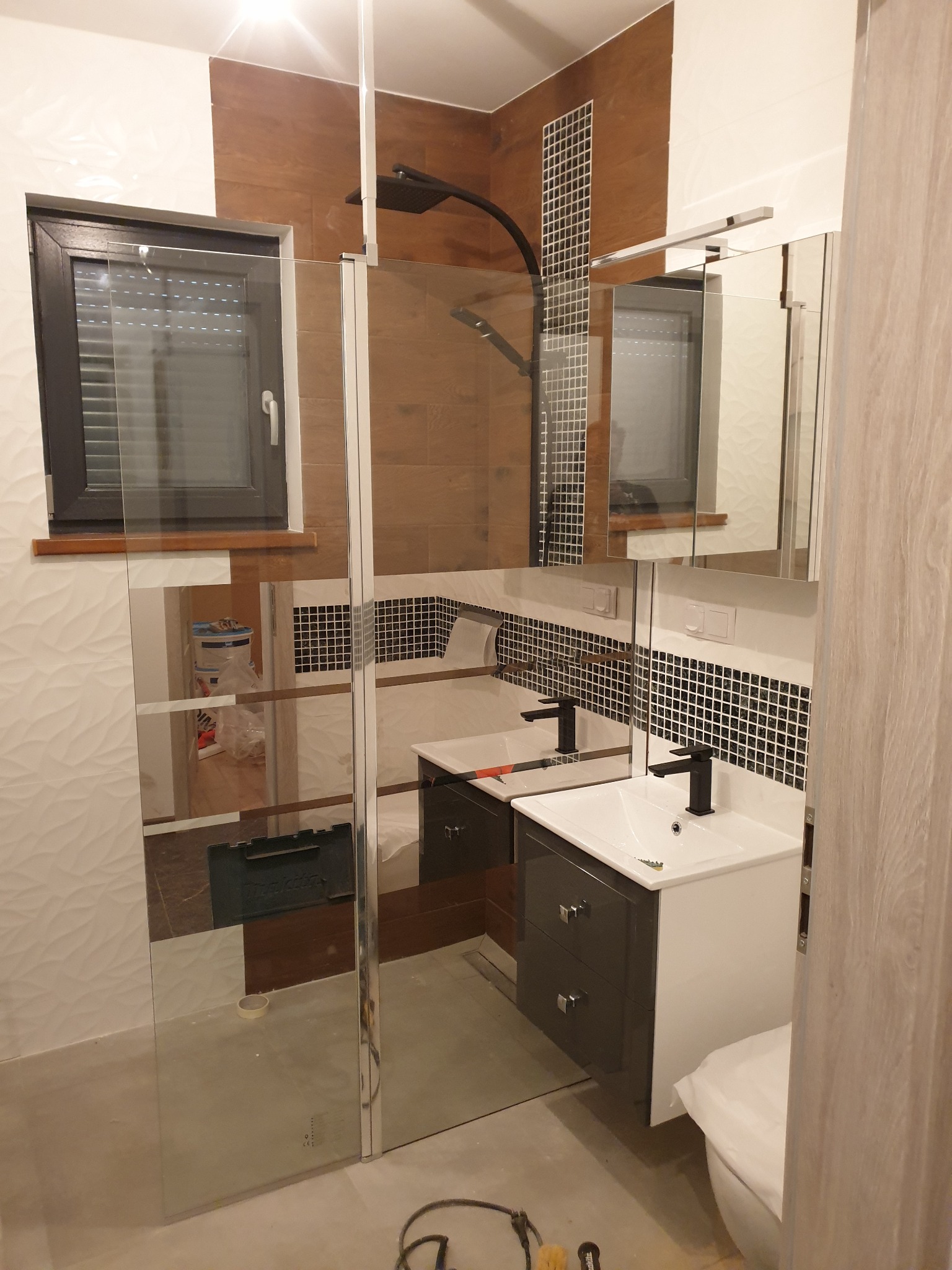 Nowoczesna łazienka z kabiną prysznicową typu walk-in, umywalką z szafką w kolorze grafitowym i białym oraz mozaiką na ścianie, widoczne narzędzia i materiały sugerują prace wykończeniowe.