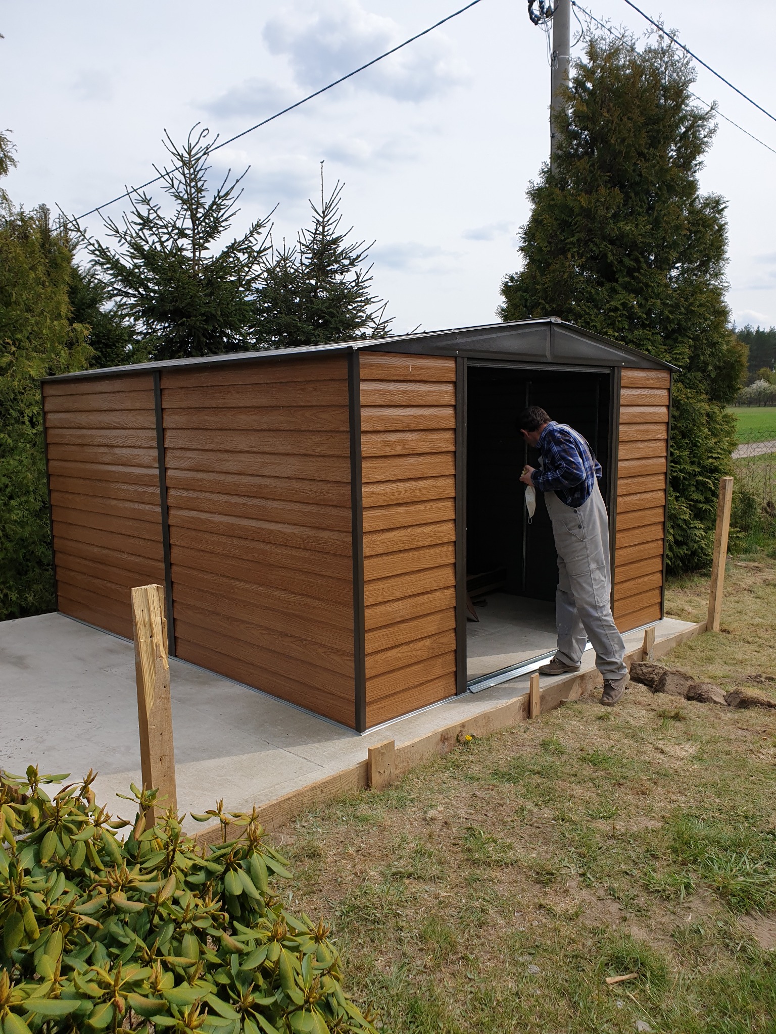 Montaż brązowego, metalowego domku narzędziowego na betonowej wylewce. Mężczyzna w szarym kombinezonie wchodzi do środka, trzymając w ręku maseczkę. Widoczne drewniane elementy szalunku. W tle drzewa.