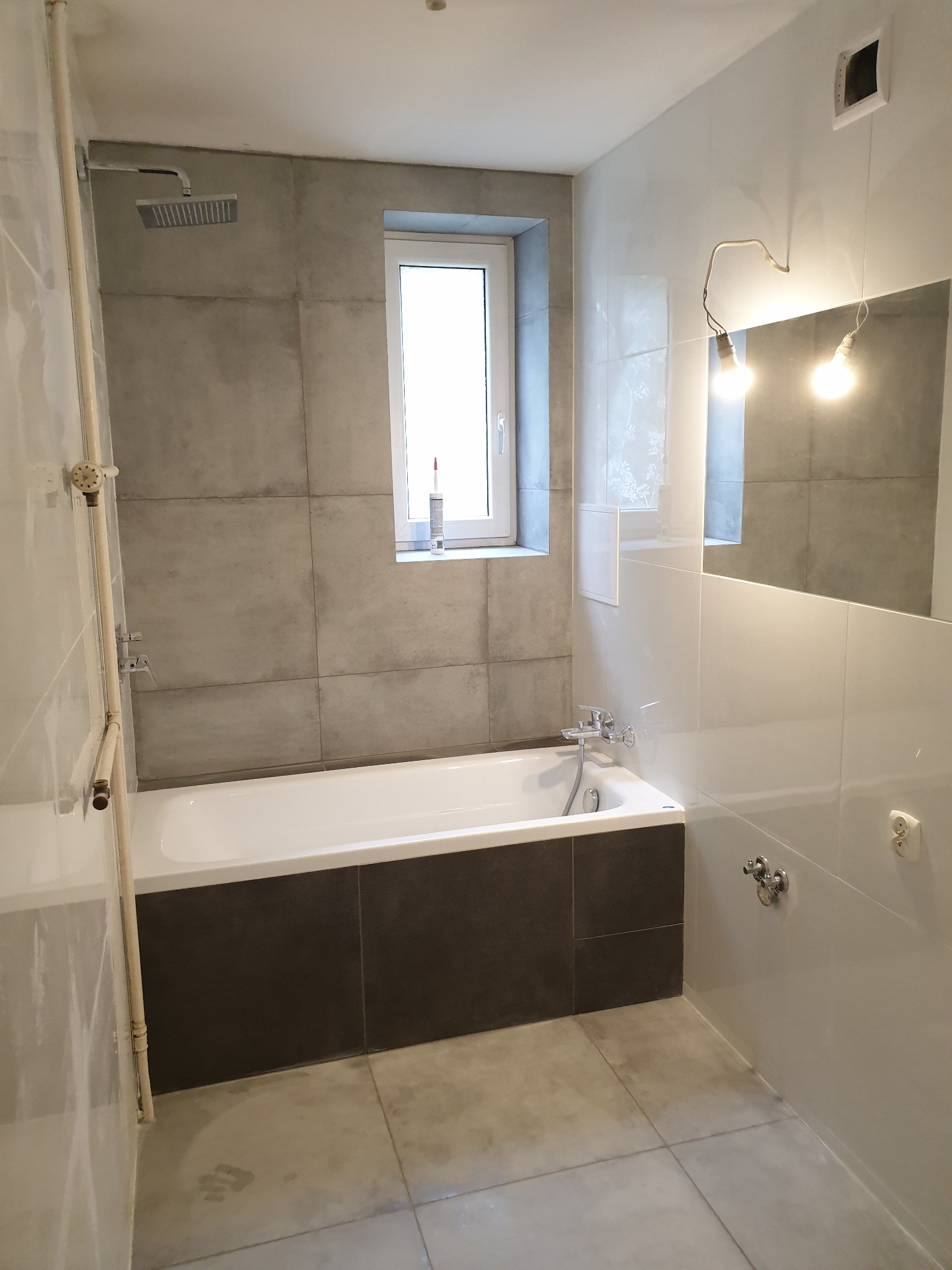 Wnętrze łazienki z wanną obudowaną ciemnymi płytkami, szare płytki na ścianach i podłodze, okno z tubą silikonu na parapecie, widoczne instalacje wodne i prowizoryczne oświetlenie.