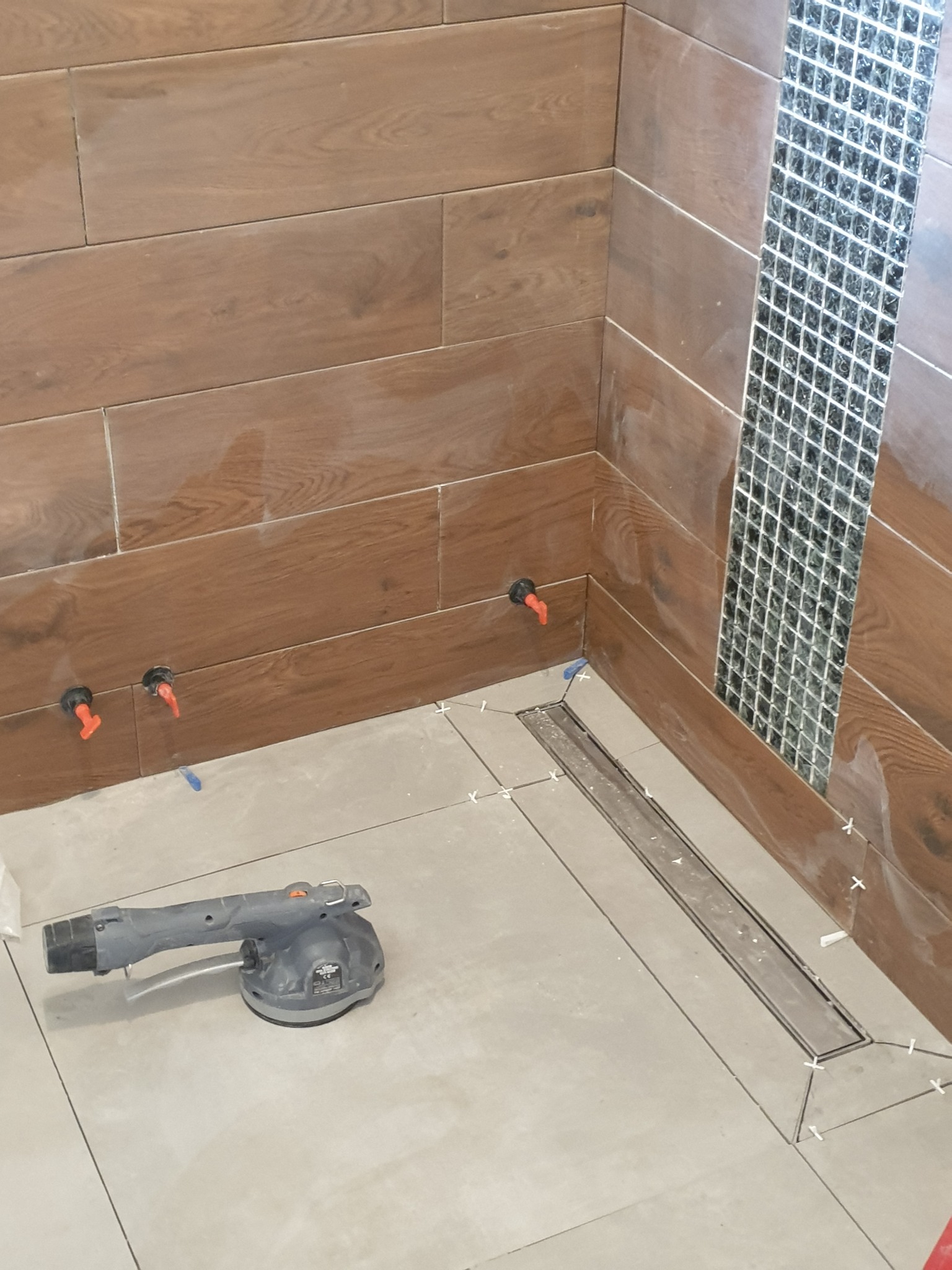 Montaż płytek drewnopodobnych na ścianie z dekoracyjnym pasem mozaiki szklanej, widoczny odpływ liniowy w posadzce i przyssawka do płytek.