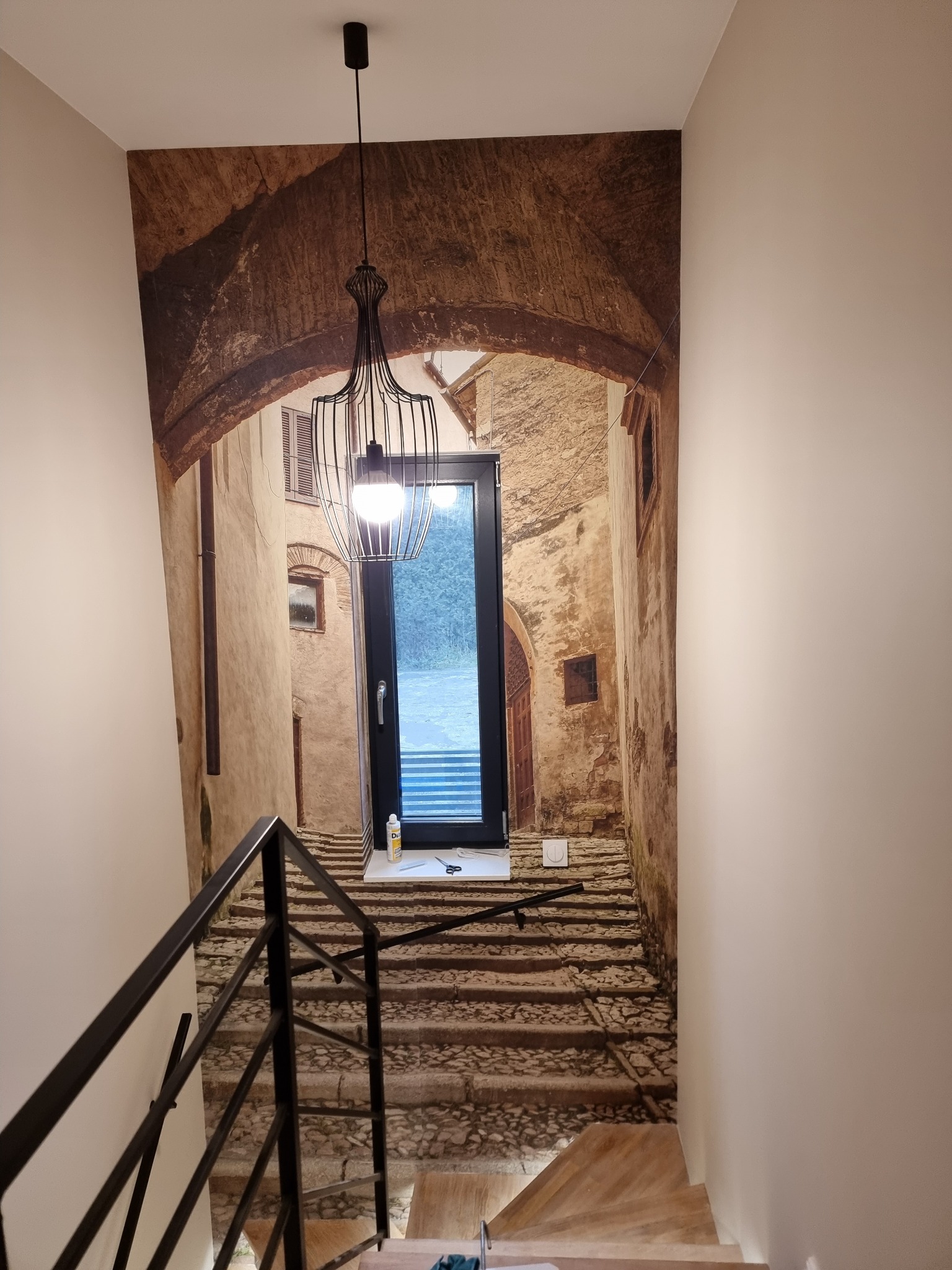Schody z czarną metalową balustradą prowadzące w dół, z fototapetą imitującą uliczkę starego miasta z łukiem i oknem, nad nimi wisi czarna lampa w stylu loft.