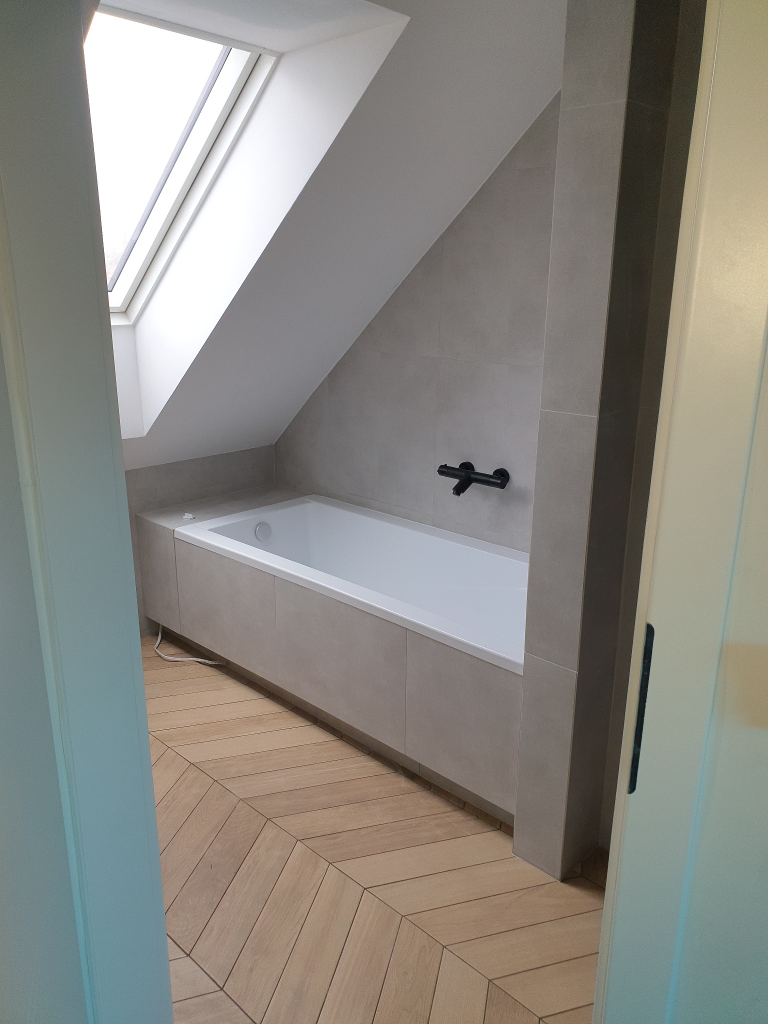 Nowoczesna łazienka na poddaszu z białą wanną zabudowaną w szarą obudowę, szare płytki ścienne, czarna bateria wannowa i parkiet ułożony we wzór jodełki francuskiej, doświetlona oknem dachowym.