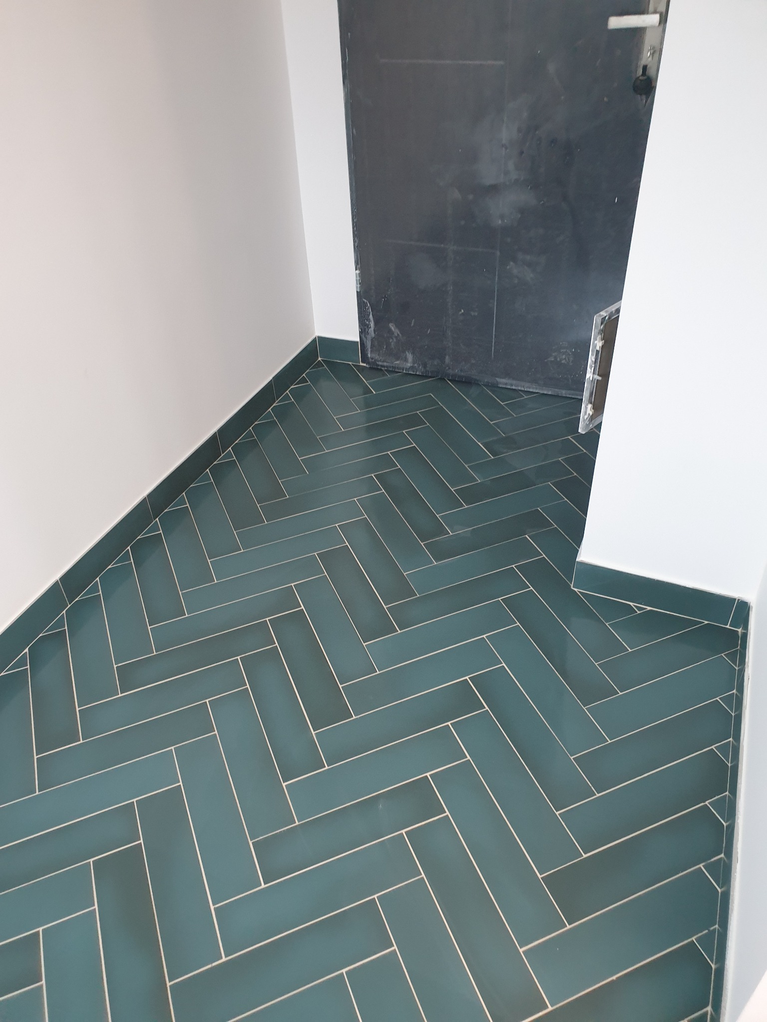 Turkusowa podłoga z płytek ułożonych w jodełkę, kontrastująca z białymi ścianami i ciemnymi drzwiami, świeżo po położeniu fugi.