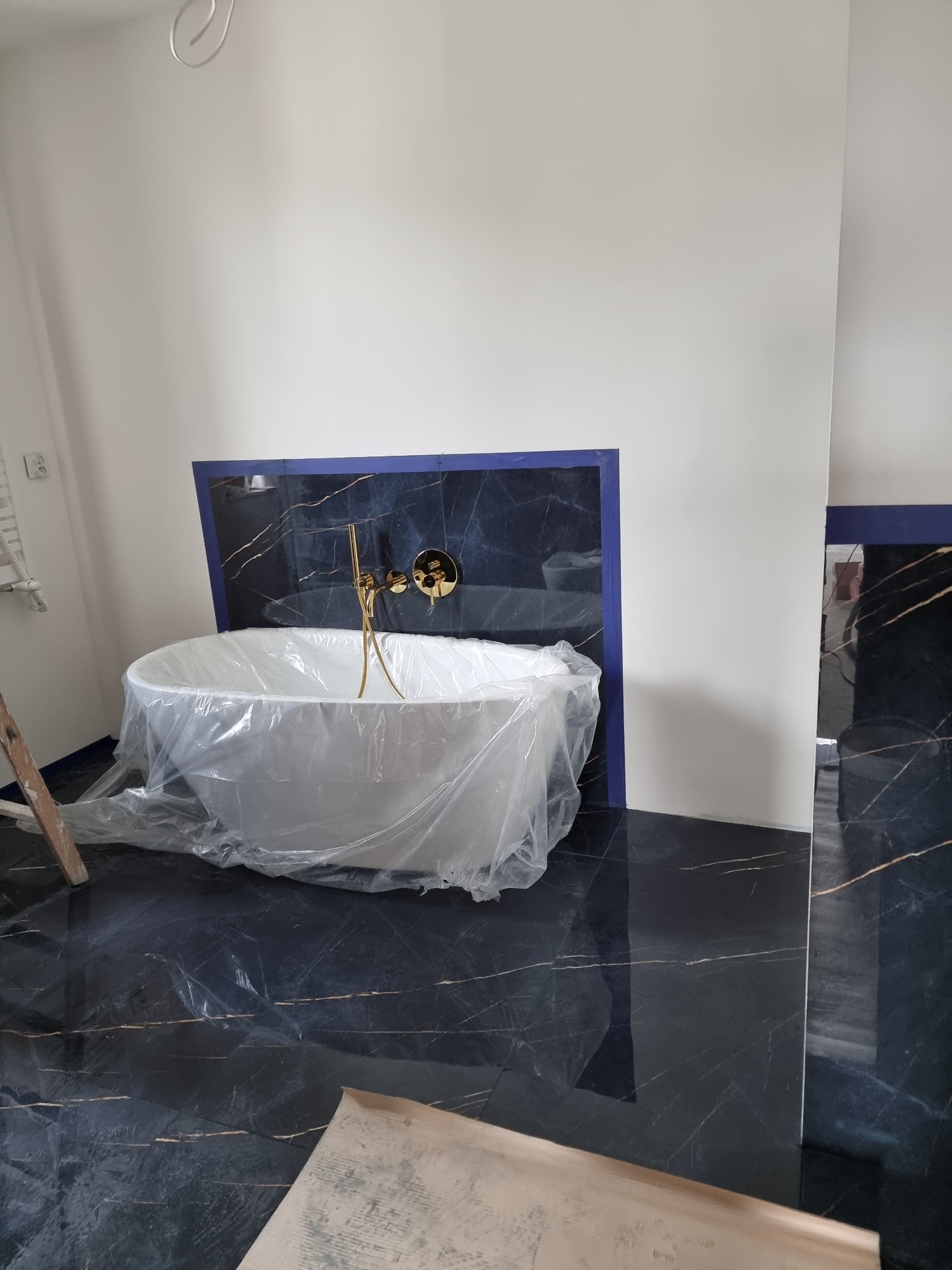 Wolnostojąca wanna owinięta folią ochronną, złota bateria zamontowana na tle ściany wyłożonej czarnym marmurem z niebieską ramą, podłoga z czarnego marmuru, drabina i kartony w tle.