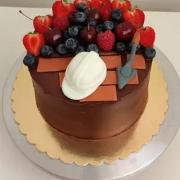 Tort czekoladowy udekorowany owocami, czekoladowymi cegłami, kaskiem i kielnią z lukru, na złotej tacy.