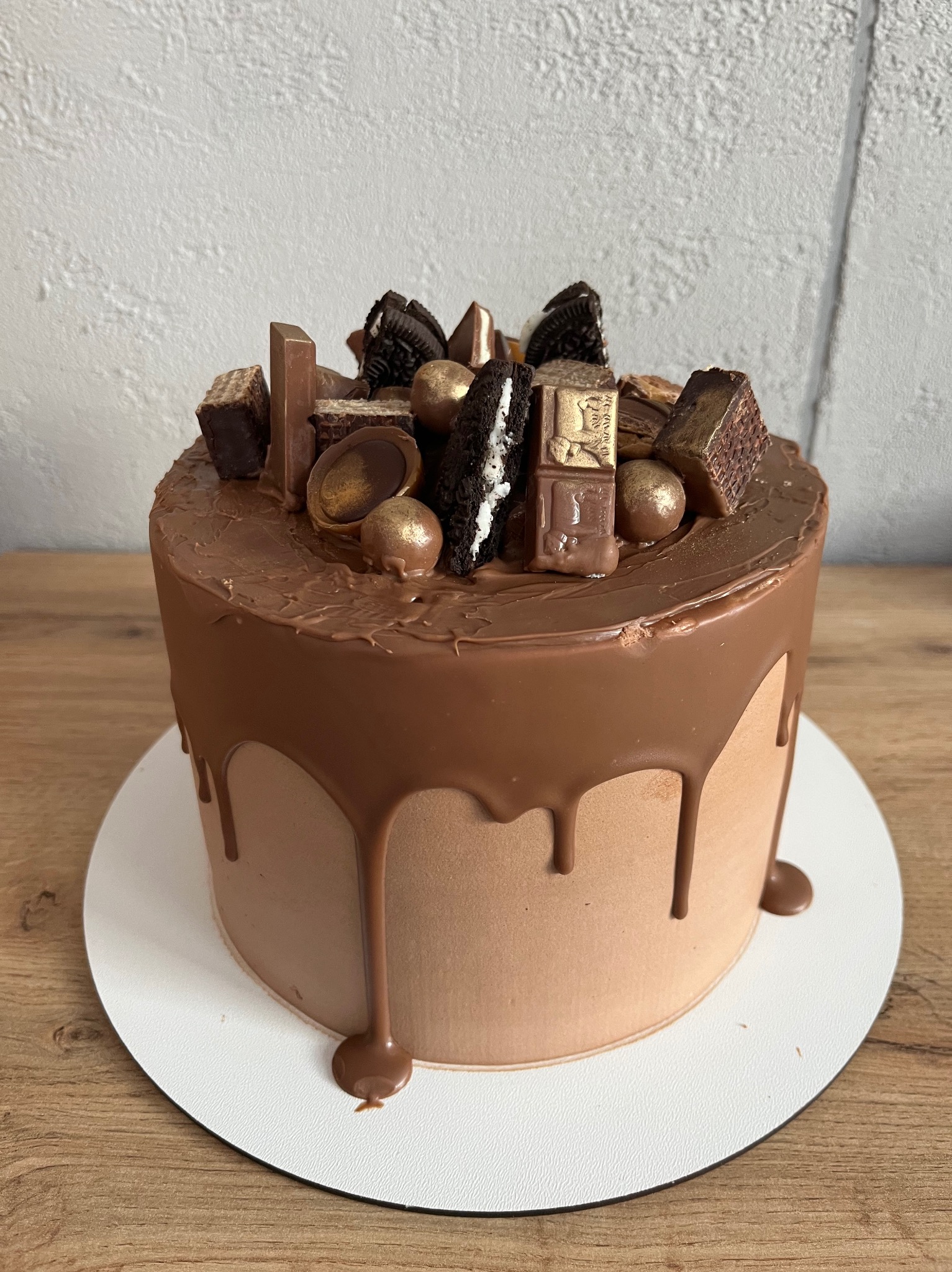 Czekoladowy tort z polewą i dekoracjami z batonów, ciastek Oreo i złotych kulek, umieszczony na białym podkładzie na drewnianym stole.