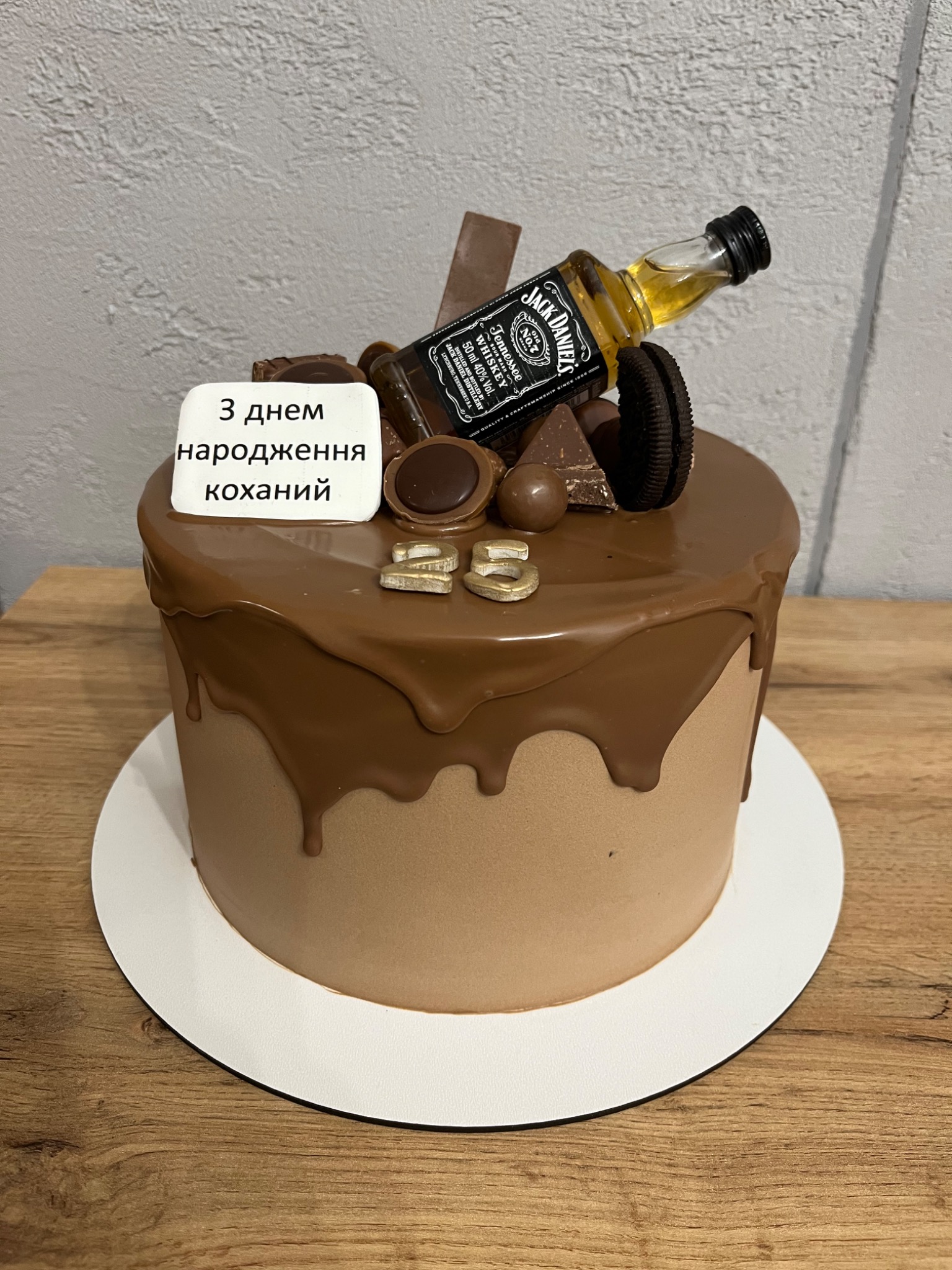 Tort urodzinowy z czekoladowym polewem, dekorowany mini butelką Jack Daniel's, czekoladkami, ciasteczkami Oreo i napisem 'З днем народження коханий' oraz cyfrą '25' w złotym kolorze.
