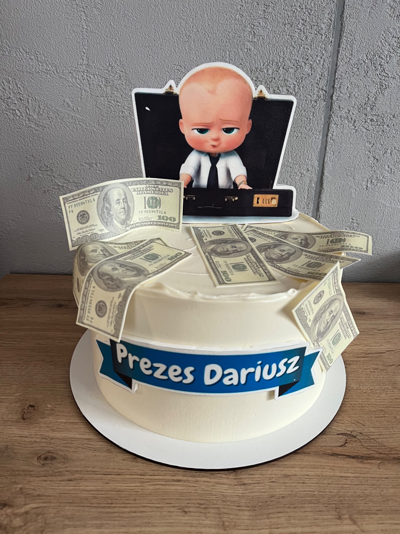 Tort urodzinowy z motywem Boss Baby, udekorowany banknotami studolarowymi i napisem 'Prezes Dariusz' na niebieskim banerze, umieszczony na drewnianym stole.