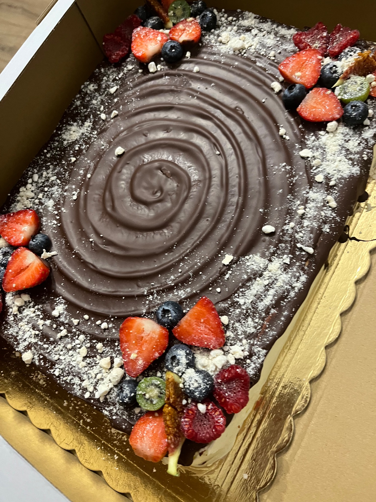 Prostokątny tort czekoladowy udekorowany świeżymi truskawkami, borówkami, malinami, kiwi oraz spiralną polewą czekoladową i posypką z białego proszku, umieszczony w złotym kartonowym pudełku.