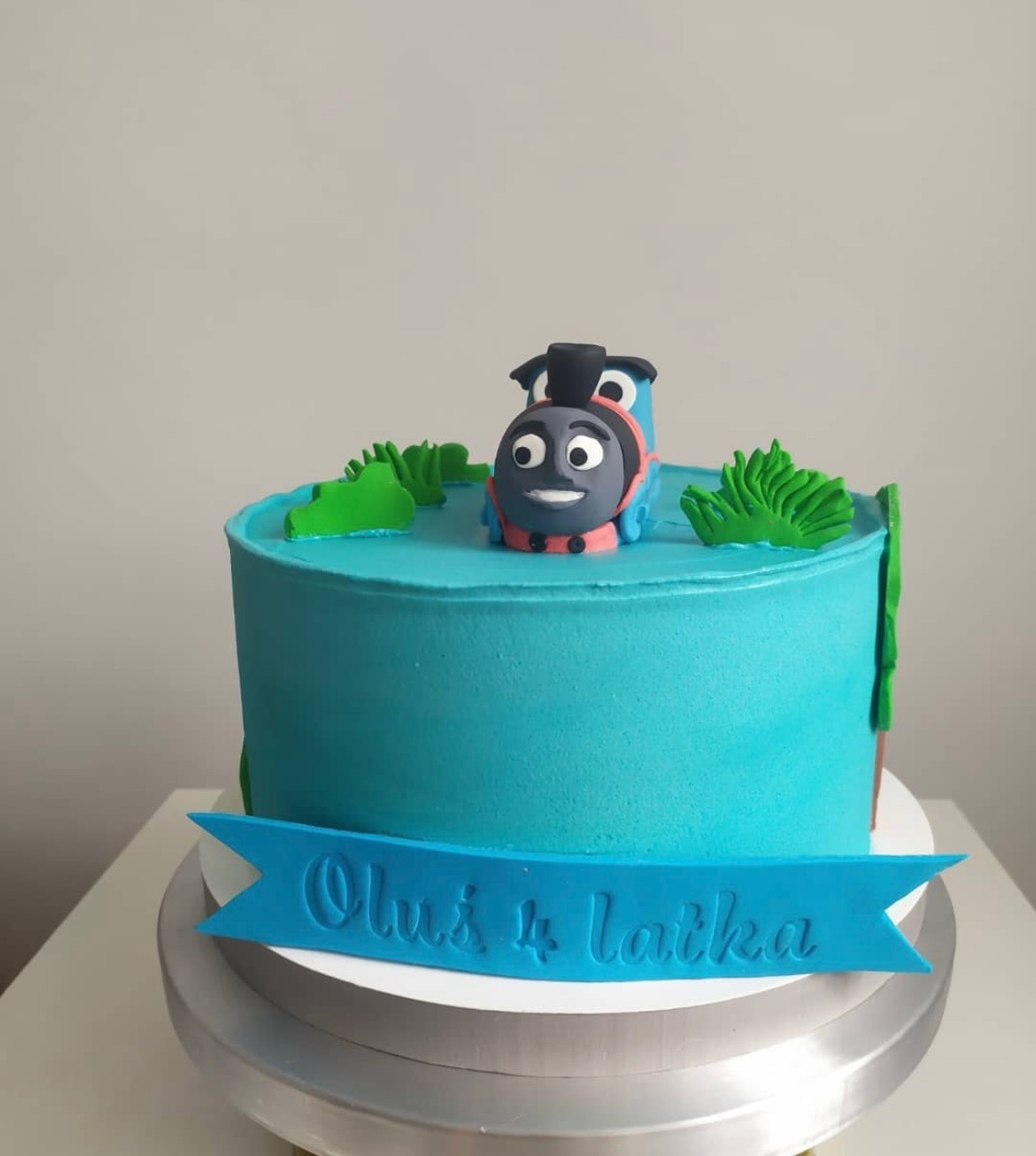 Tort urodzinowy z turkusowym lukrem, dekorowany figurką lokomotywy Tomek i zielonymi elementami roślinnymi, z napisem 'Oluś 4 latka' na niebieskiej wstążce.