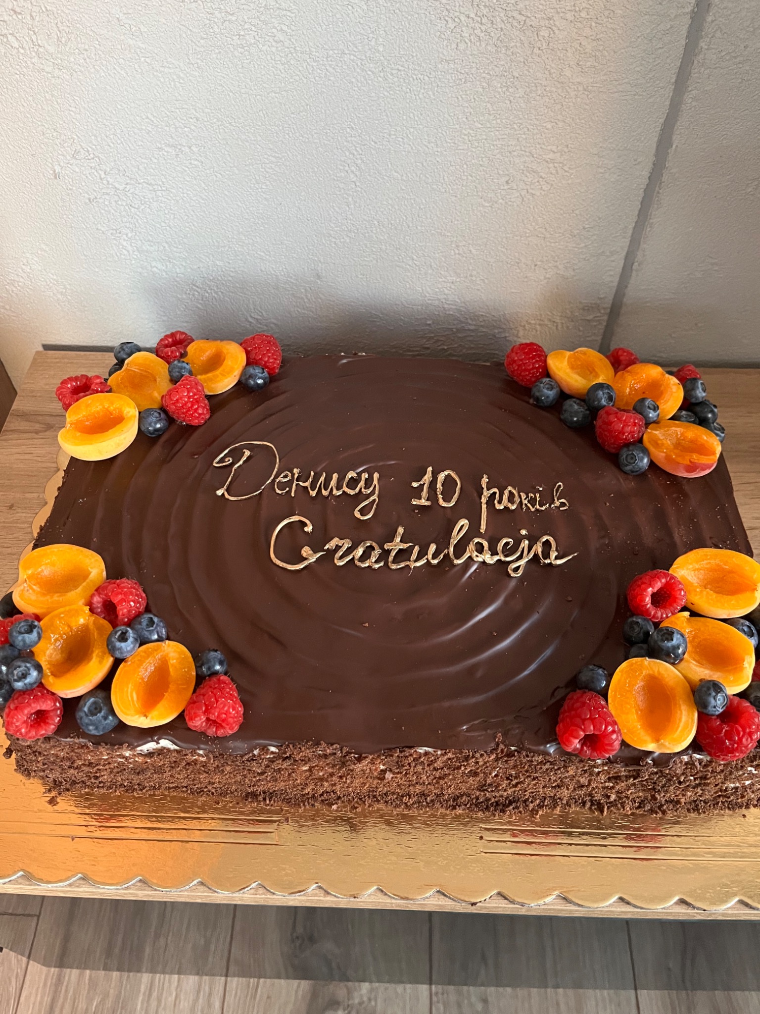 Prostokątny tort czekoladowy z napisem 'Denucy 10 років Gratulaćja' wykonanym złotym lukrem, udekorowany po bokach połówkami moreli, malinami i borówkami, umieszczony na złotej podkładce.