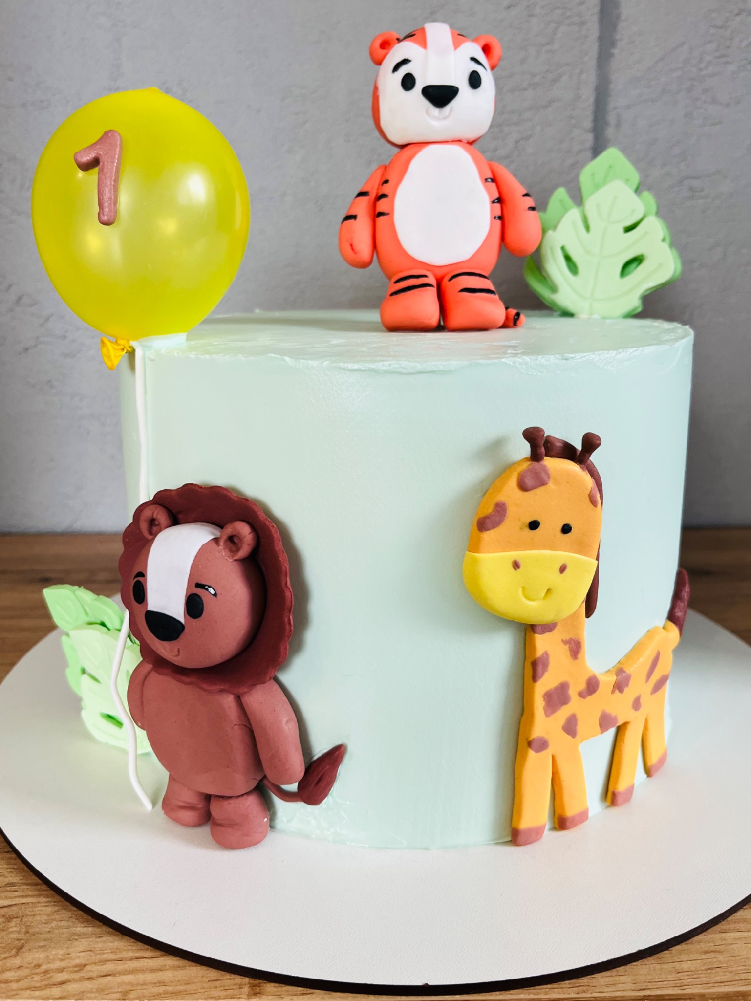 Udekorowany tort urodzinowy z motywem safari z Żor, z figurkami lwa, żyrafy, tygrysa i balonem z numerem jeden, na gładkim, jasnym tle.