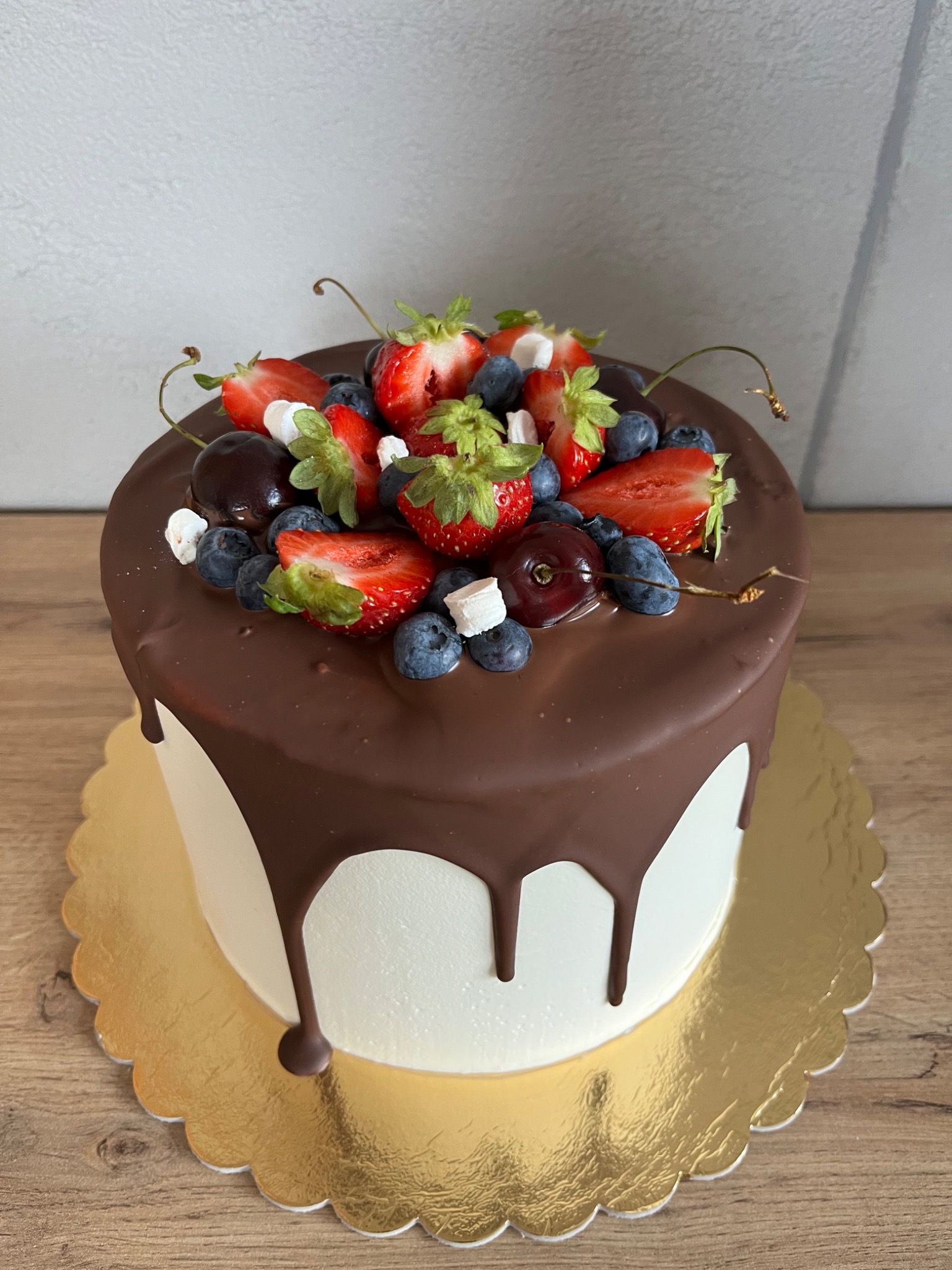 Tort z polewą czekoladową, udekorowany świeżymi truskawkami, borówkami, wiśniami i małymi kostkami pianek, umieszczony na złotej podkładce na drewnianym blacie.