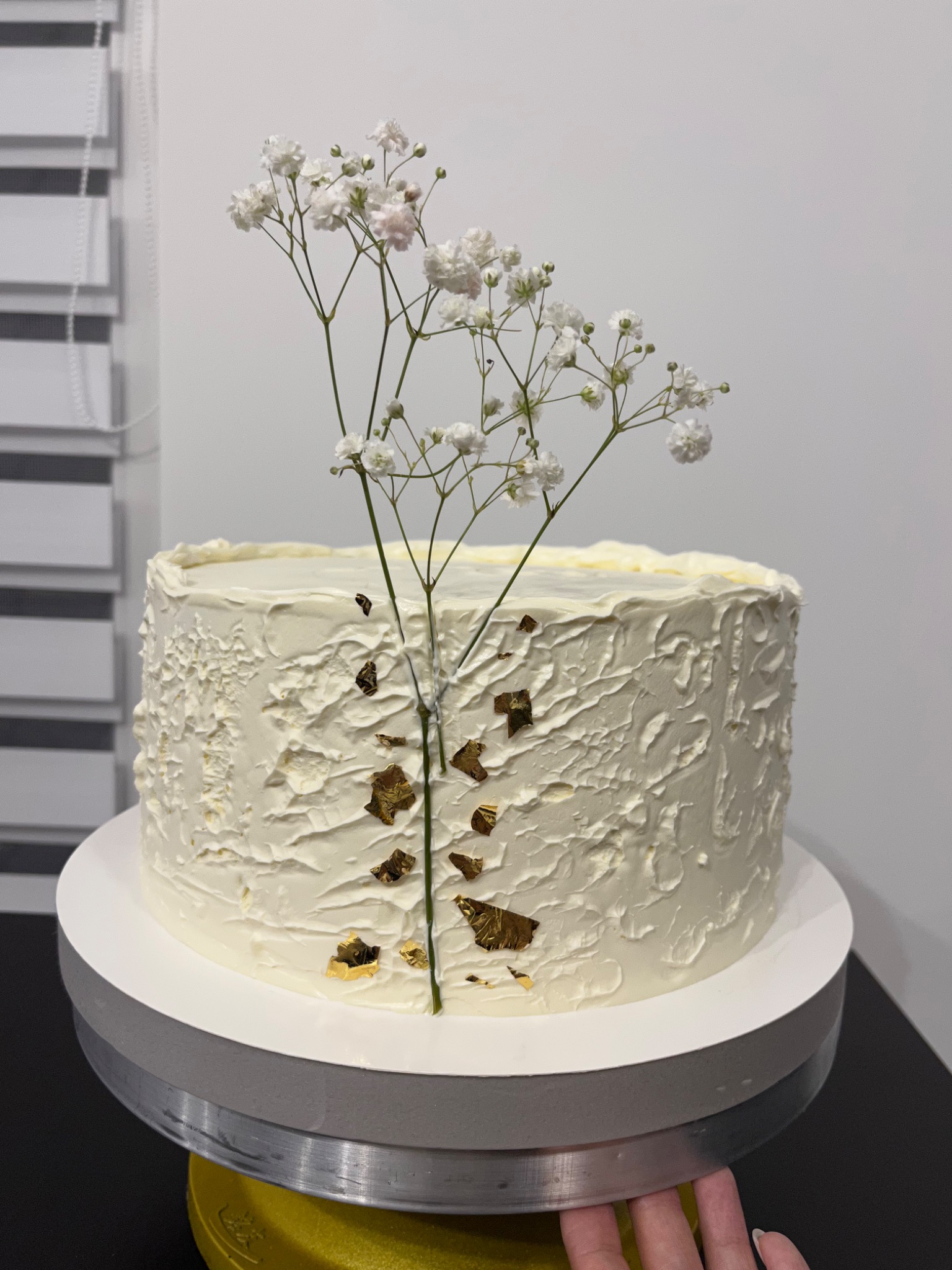 Tort z białym kremem o nieregularnej fakturze, ozdobiony gipsówką i płatkami złota, umieszczony na obrotowym stojaku.