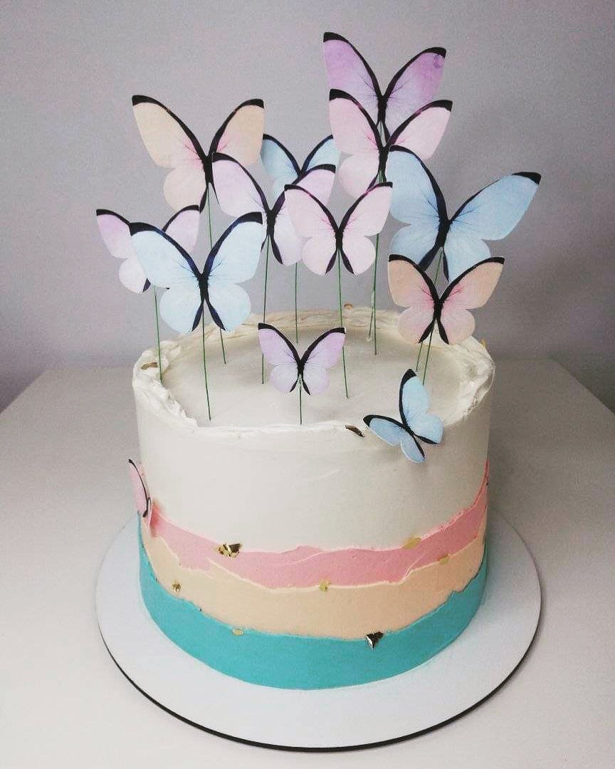 Tort urodzinowy z białym kremem, pastelowymi warstwami i motylkami na drucikach, dekorowany płatkami złota.