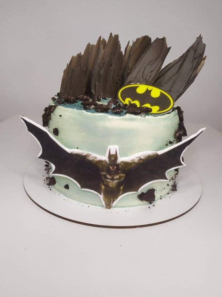 Tort z motywem Batmana: jasnoniebieski krem, czekoladowe elementy dekoracyjne, logo Batmana i figurka postaci z rozpostartymi skrzydłami.