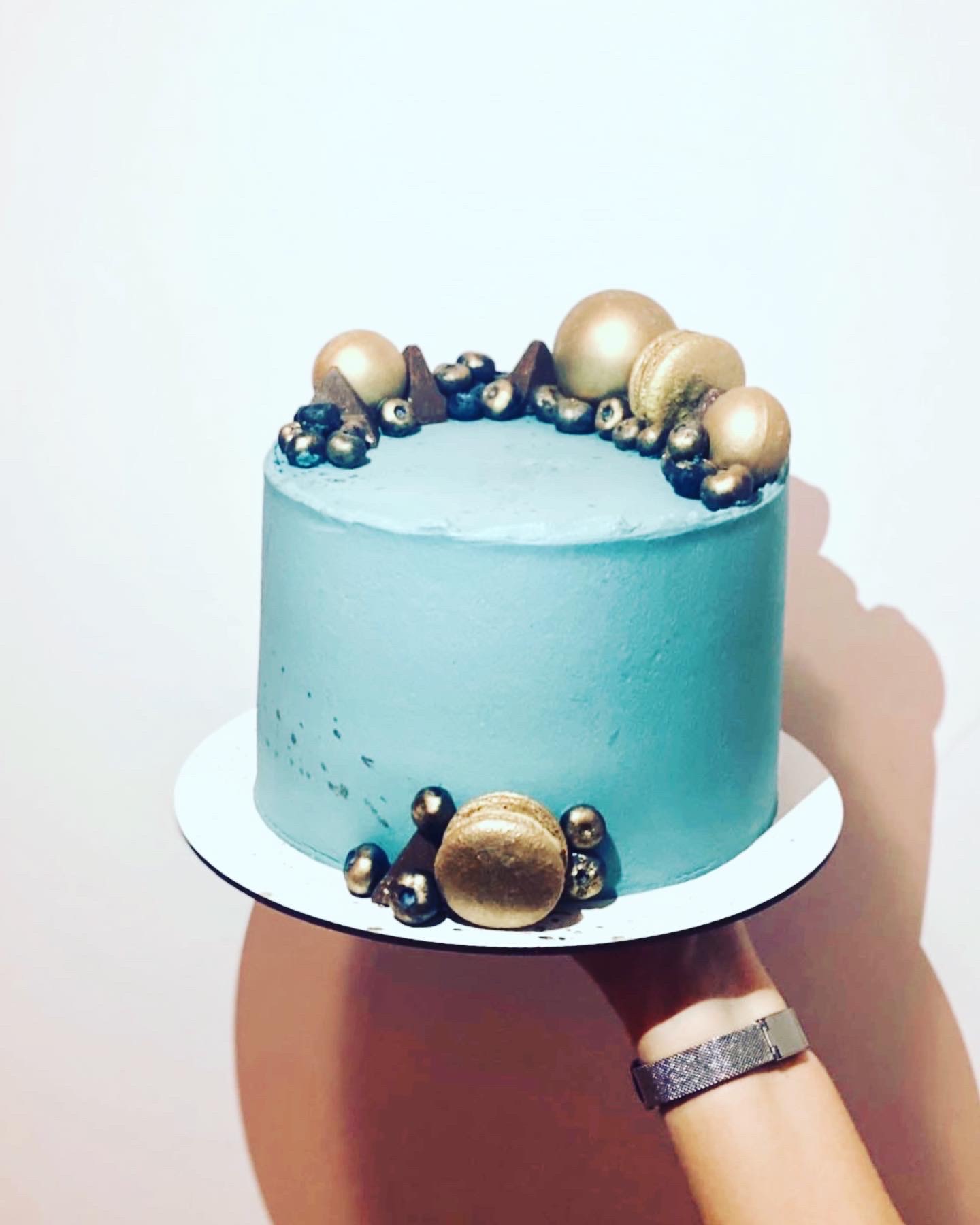 Niebieski tort z dekoracjami z złotych kulek, makaroników i czekoladowych trójkątów, trzymany na białym talerzu przez osobę z srebrną bransoletką.
