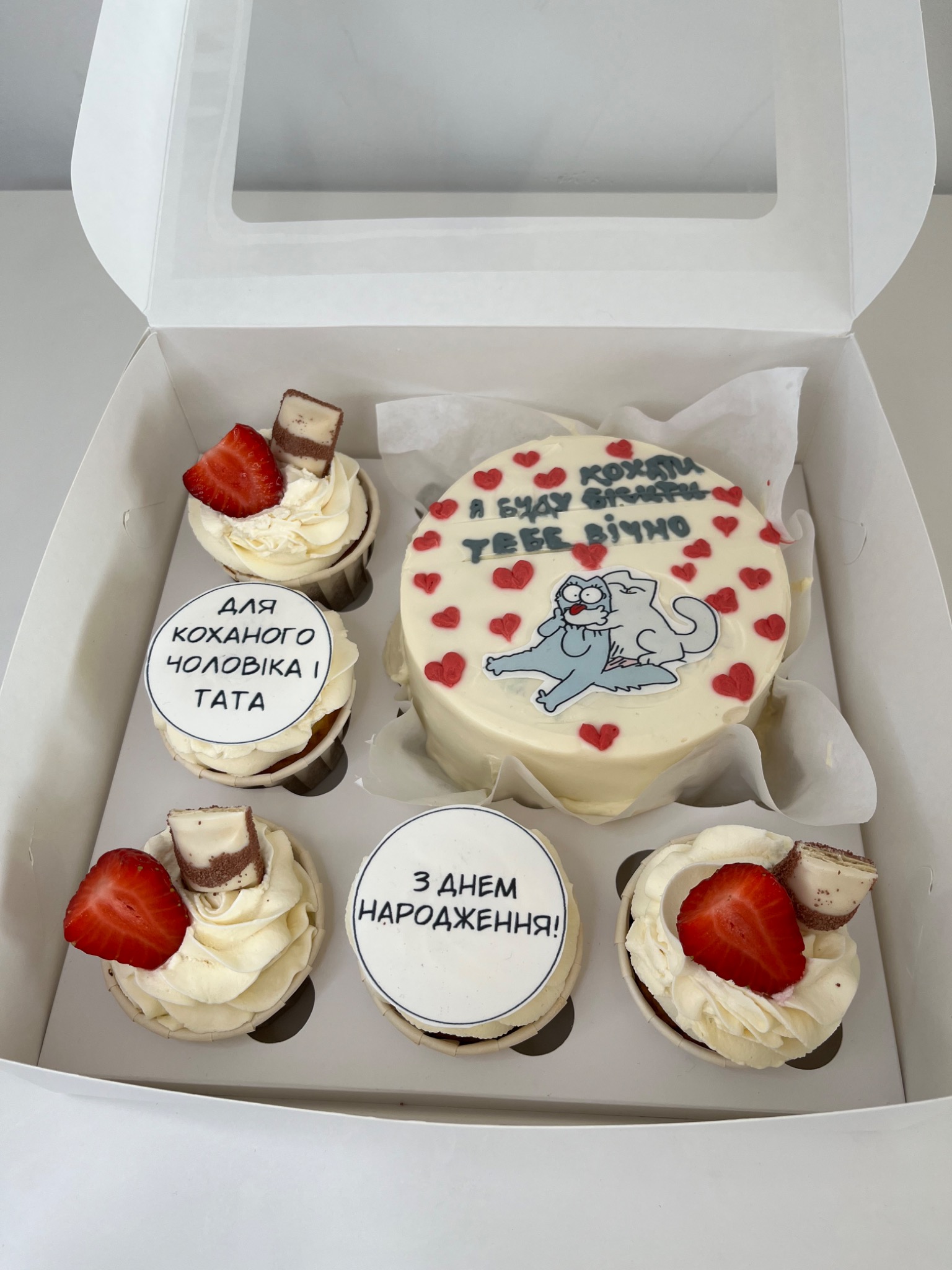Ozdobny zestaw słodkości w białym pudełku: tort z napisem i rysunkiem kota, otoczony babeczkami z kremem, truskawkami i kawałkami czekolady. Na babeczkach okrągłe dekoracje z napisami.