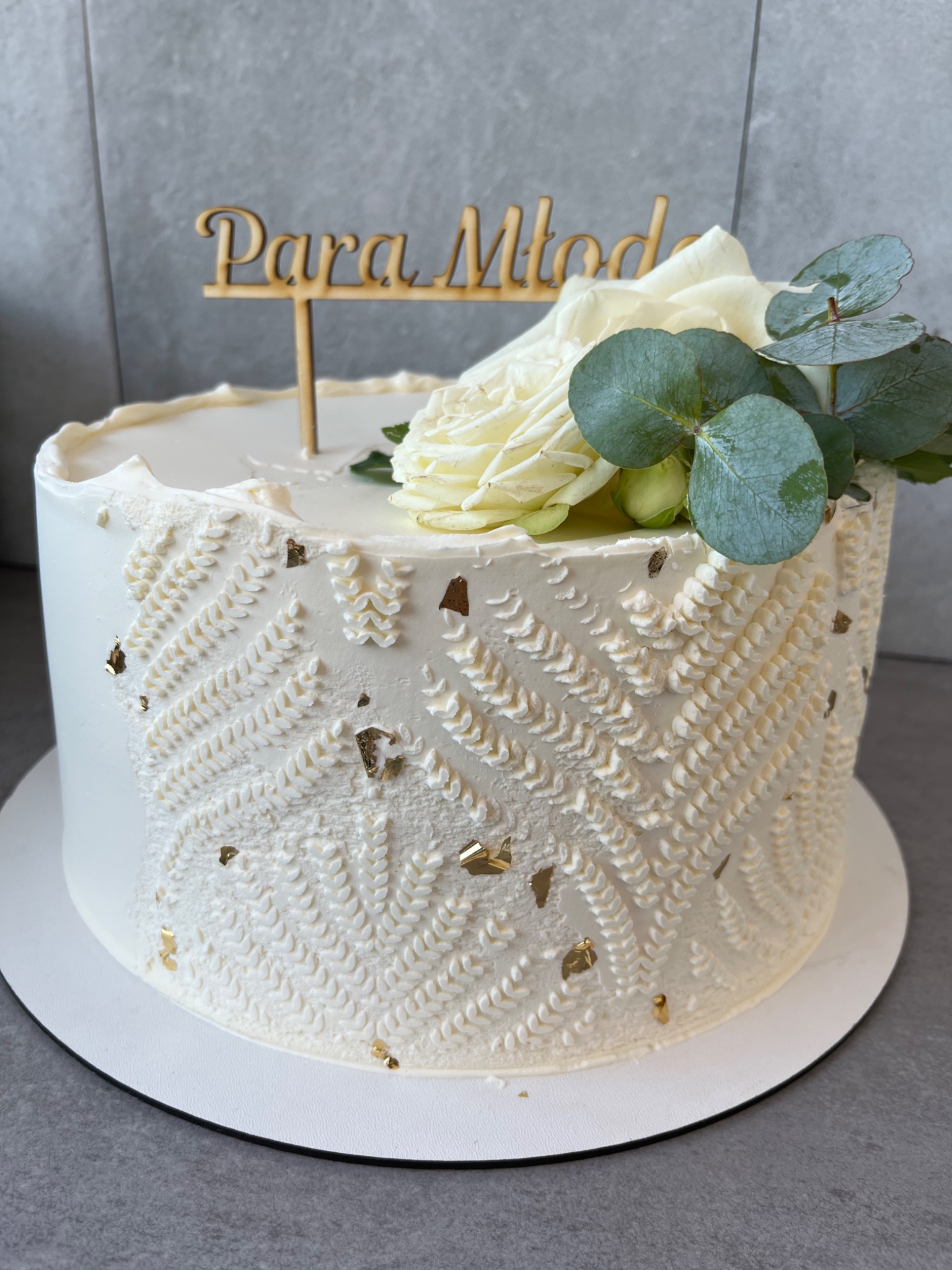 Tort weselny z białym kremem, dekorowany wzorem przypominającym dziergany sweter, złotymi płatkami i napisem 'Para Młoda' oraz białą różą i gałązką eukaliptusa.