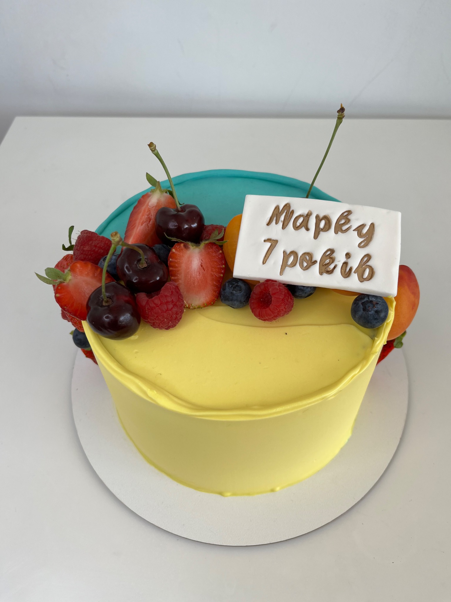 Tort urodzinowy z żółtym kremem, udekorowany świeżymi truskawkami, wiśniami, malinami i borówkami. Na górze biała tabliczka z napisem w języku ukraińskim 'Марку 7 років' (Markowi 7 lat).