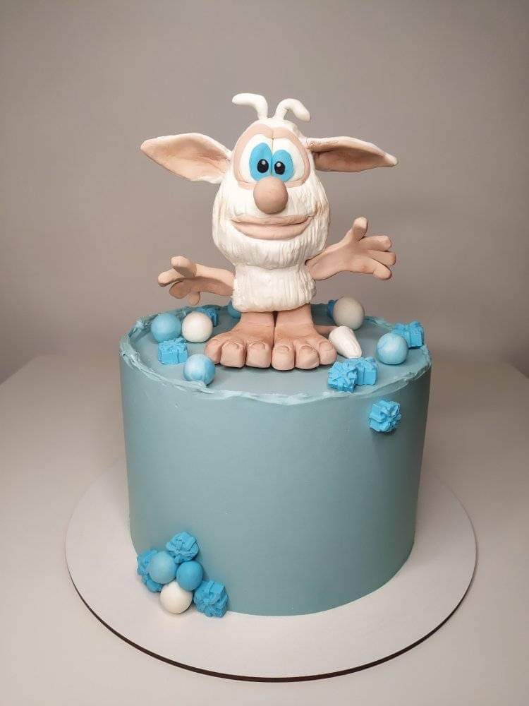 Tort w odcieniach błękitu z figurką białego stworka z dużymi oczami i uśmiechem, udekorowany kulkami.
