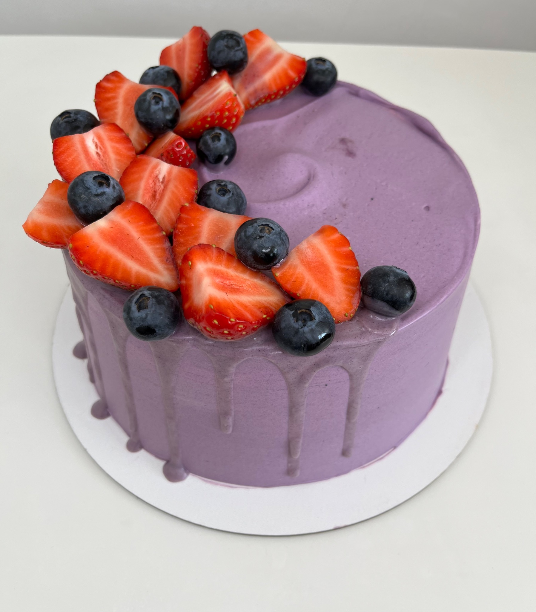 Okrągły tort z fioletowym kremem, udekorowany świeżymi truskawkami i borówkami, z kremem spływającym po bokach.