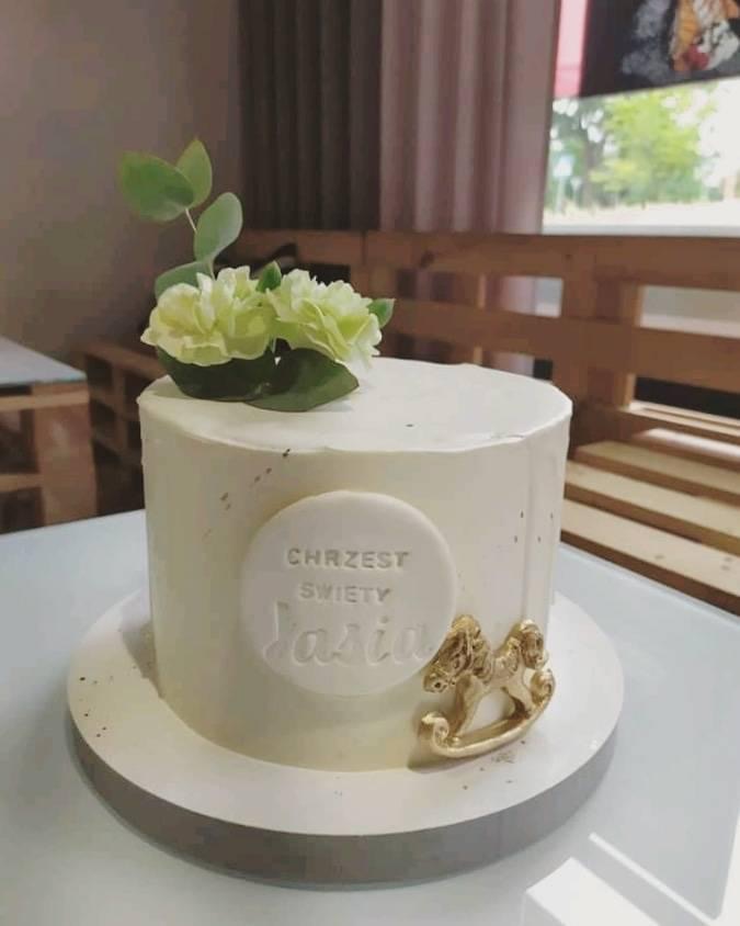 Tort na chrzest z białym lukrem, dekorowany kwiatami, listkami eukaliptusa, napisem 'Chrzest Święty' i złotym konikiem na biegunach.