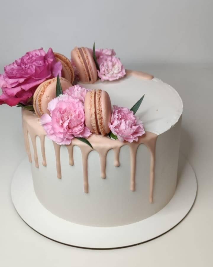 Elegancki tort z białym kremem, różowymi dripami, makaronikami i goździkami na wierzchu, umieszczony na białym podeście.