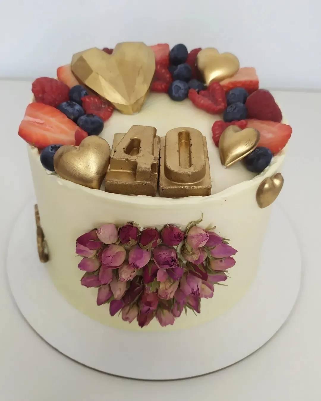 Tort urodzinowy z kremem, dekorowany złotymi sercami, owocami (truskawki, borówki, maliny) i suszonymi pączkami róż ułożonymi w kształt serca, z napisem '40' z czekolady.