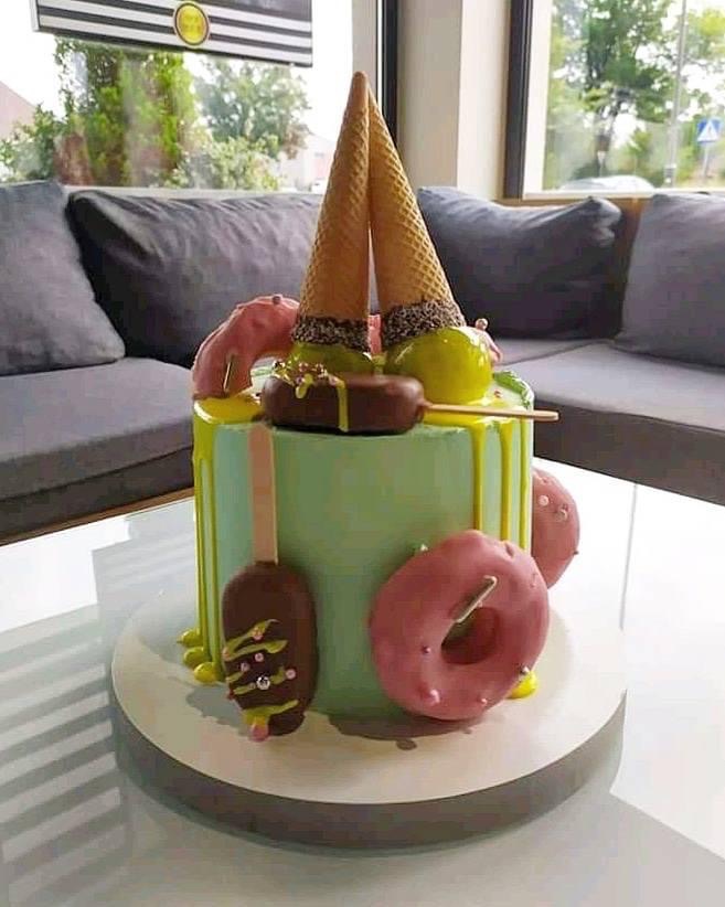 Nietypowy tort urodzinowy z zielonym lukrem, dekorowany lodami w waflach, lizakami czekoladowymi i różowymi pączkami, ustawiony na szklanym stole w jasnym wnętrzu.