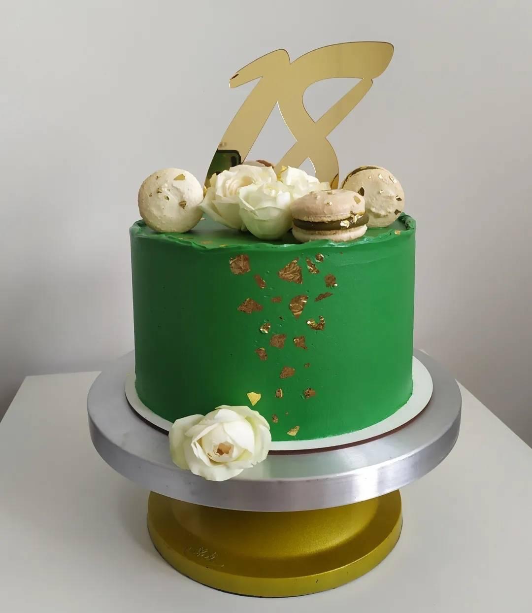 Zielony tort urodzinowy z dekoracją z białych róż, makaroników i złotych płatków, na metalowej podstawie obrotowej.