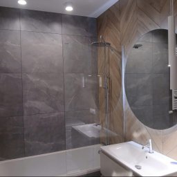 WYREMONTOWANI.COM - Nowoczesna łazienka z wanną, szklanym parawanem i deszczownicą. Ściany wyłożone szarymi płytkami imitującymi marmur oraz drewnopodobnymi w jodełkę. Okrągłe lustro nad białą umywalką.