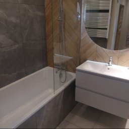 WYREMONTOWANI.COM - Nowoczesna łazienka z wanną, szklanym parawanem, podwieszaną szafką z umywalką i okrągłym lustrem. Ściany zdobią płytki imitujące ciemny marmur i jasne drewno w jodełkę.