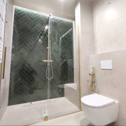 WYREMONTOWANI.COM - Kabina prysznicowa ze szklanymi drzwiami i ciemnozielonymi płytkami w jodełkę, podkreślona złotymi profilami i armaturą. Obok biały grzejnik drabinkowy i podwieszana toaleta na beżowych ścianach.