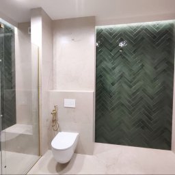 WYREMONTOWANI.COM - Elegancka łazienka z ciemnozielonymi płytkami w jodełkę, beżowymi ścianami, złotymi armaturami i podświetleniem LED akcentującym ścianę. Widoczna kabina walk-in i podwieszana toaleta.